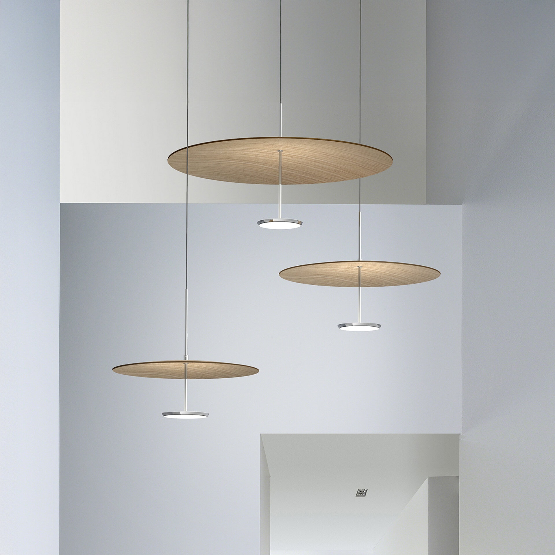 Three modern pendant lights, each with a circular wooden disc and an illuminated LED fixture, suspended at different heights in a minimalist gray and white room.