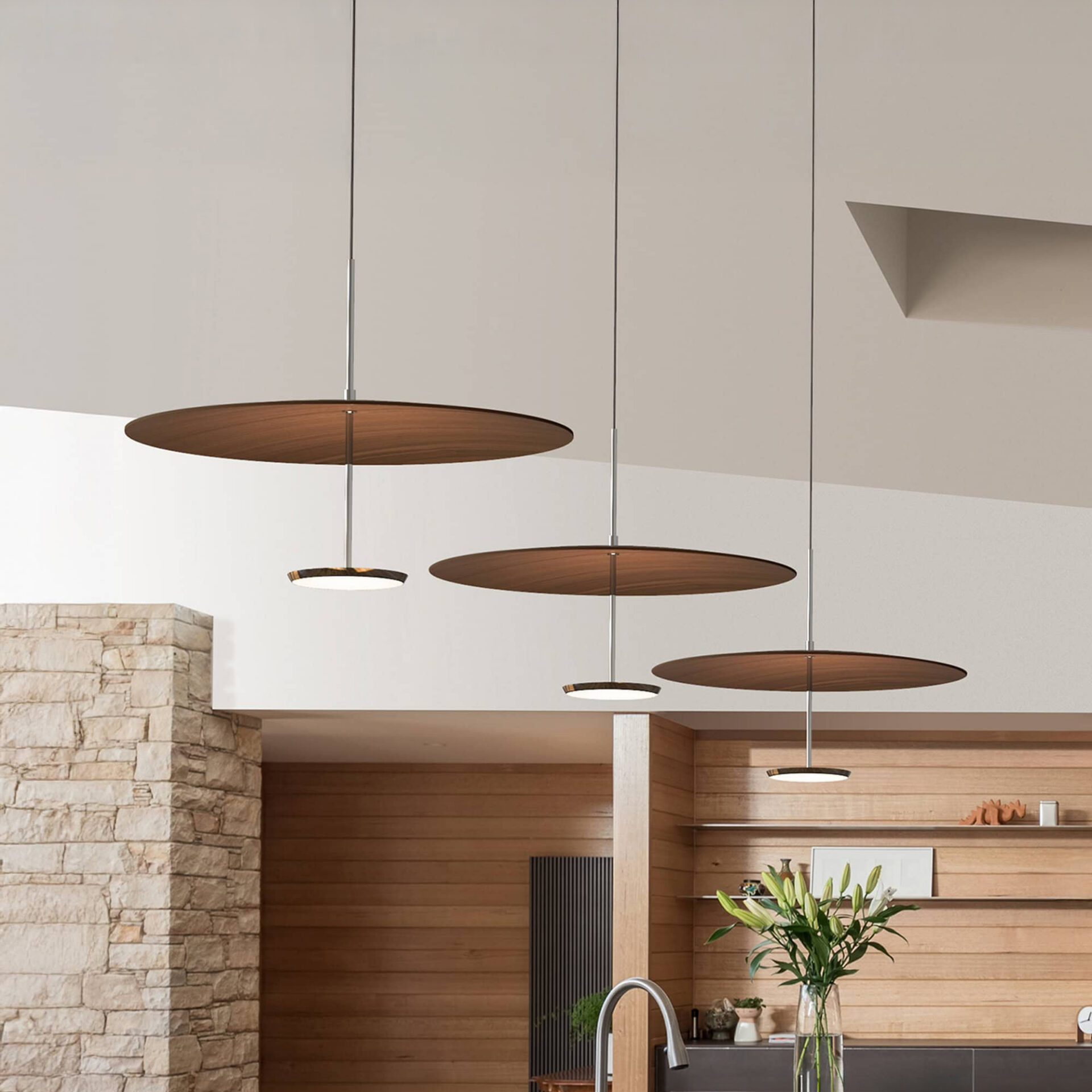 Three modern pendant lights with large wooden discs and smaller illuminated fixtures hang from a white ceiling over a kitchen island. The room features a stone pillar, horizontal wooden paneling, and a vase of white lilies.