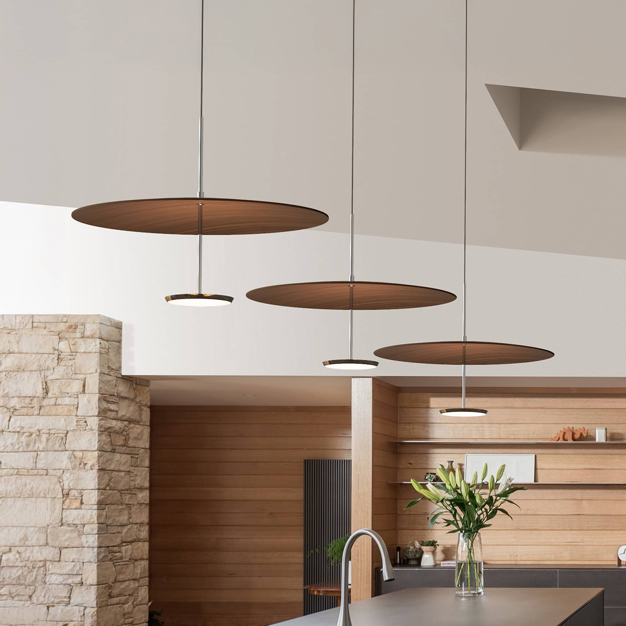 Three modern pendant lights with large wooden discs and smaller illuminated fixtures hang from a white ceiling over a kitchen island. The room features a stone pillar, horizontal wooden paneling, and a vase of white lilies.