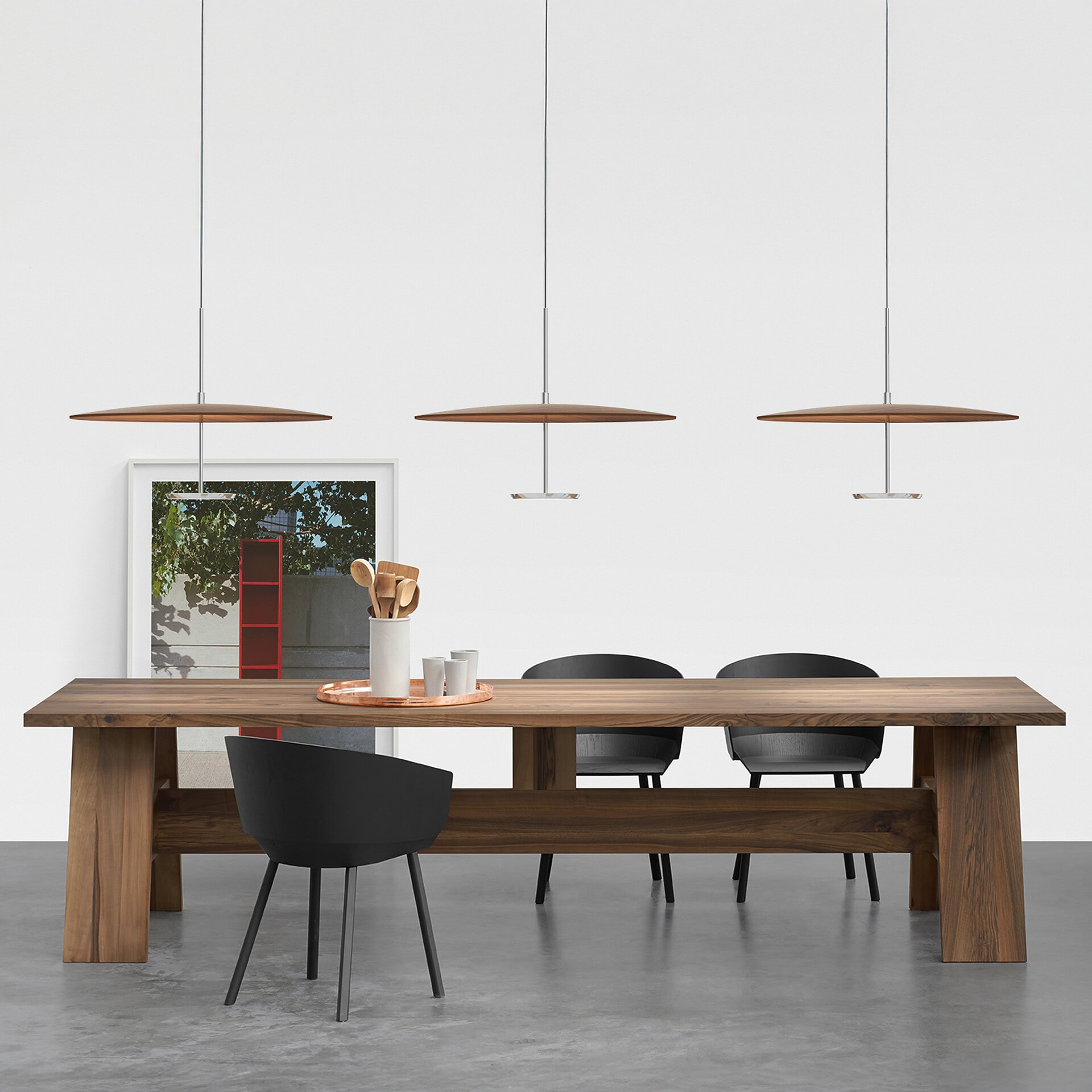 A modern dining room with a long, solid wooden table, four black chairs, and three wooden disc pendant lights. A framed artwork and kitchen utensils are on the table against a white wall and concrete floor.