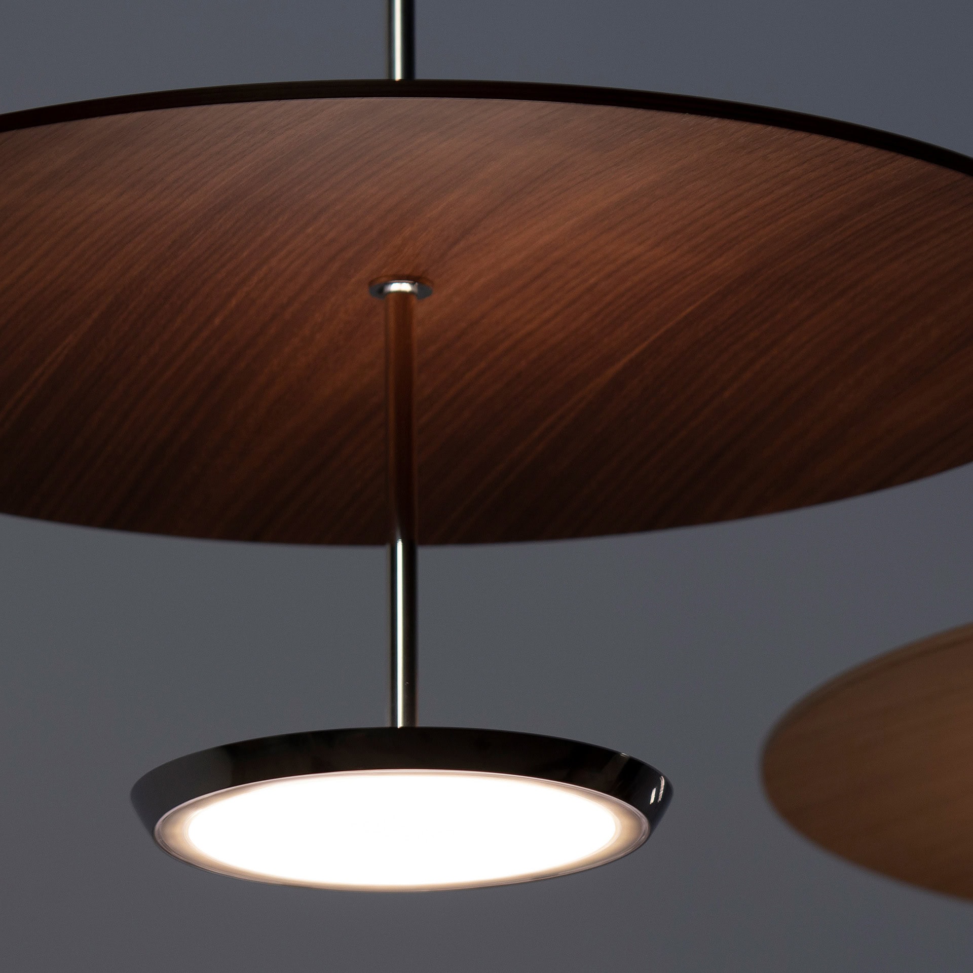A modern pendant light fixture with a dark wooden disc above a glowing circular LED light, suspended by a thin metal rod against a dark background.