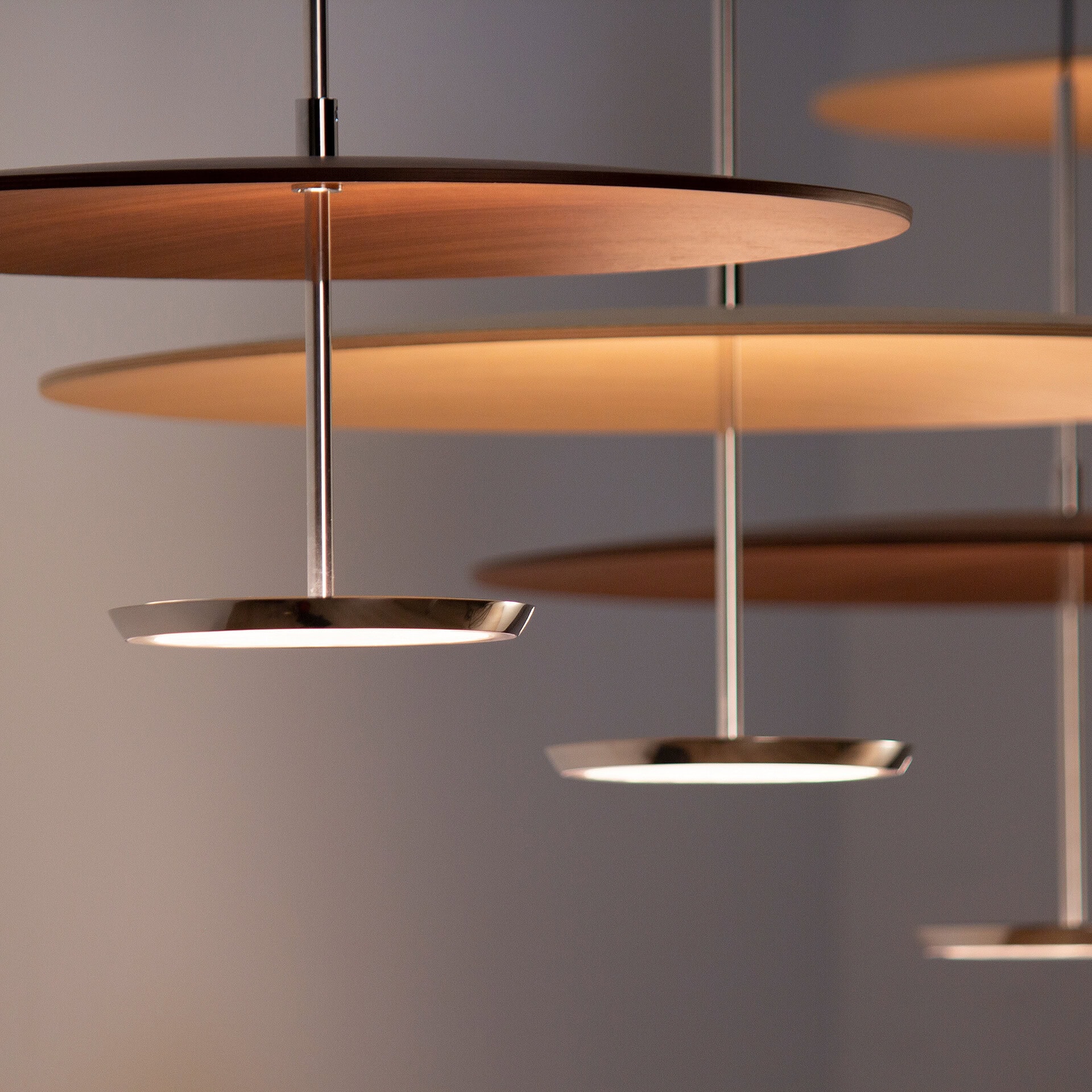 Multiple modern pendant lights with circular wooden tops and smaller, chrome LED bases, suspended against a soft gray background. The lights cast a warm glow.