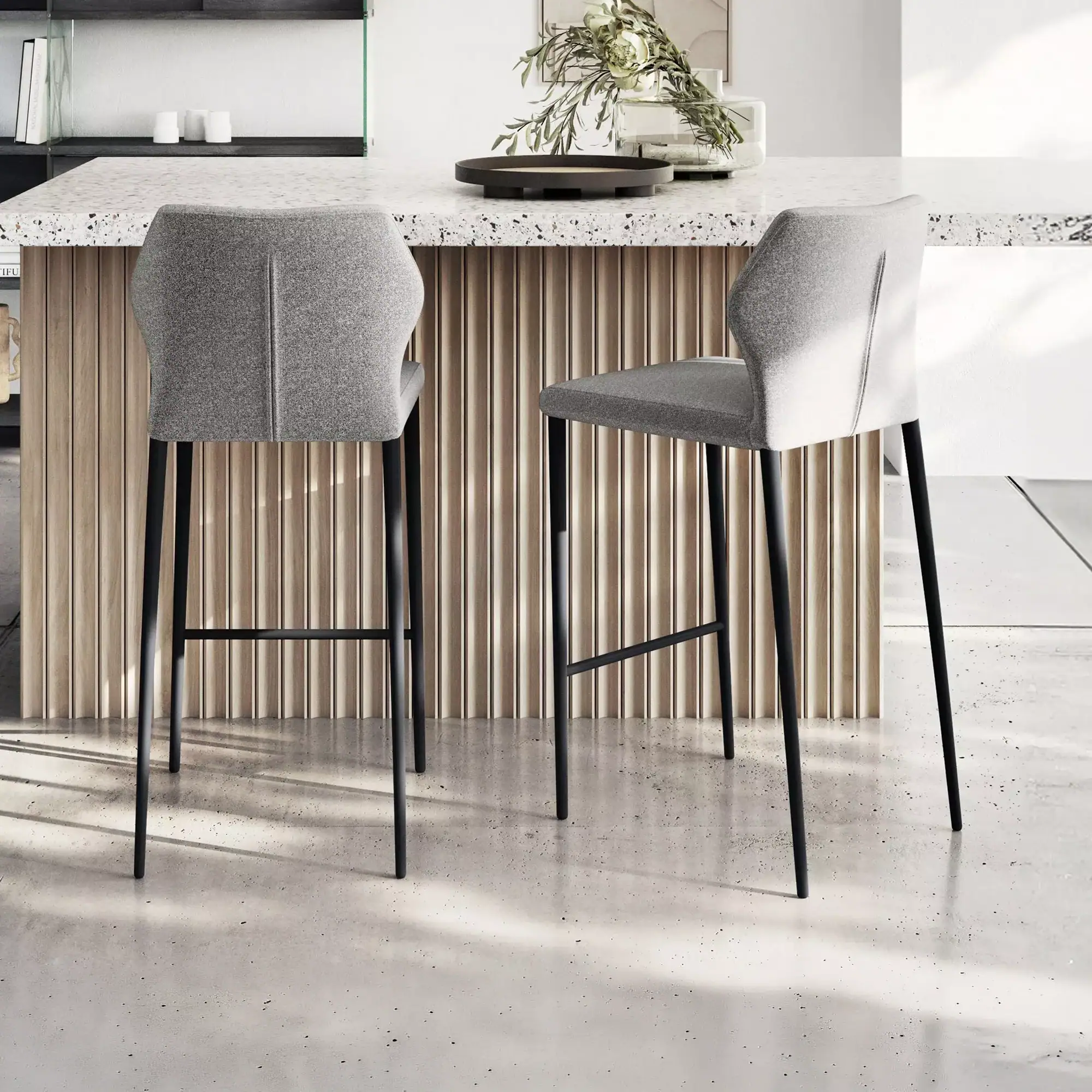 Two grey upholstered bar stools with black legs in front of a kitchen island with a terrazzo top and wooden slatted base. A vase with foliage is on the counter.