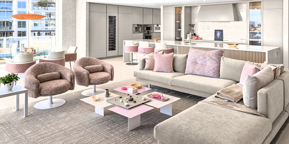 An open-concept living room, dining area, and kitchen with modern furniture in grey, white, and blush pink, featuring a sectional sofa, unique coffee tables, and a sleek kitchen island.