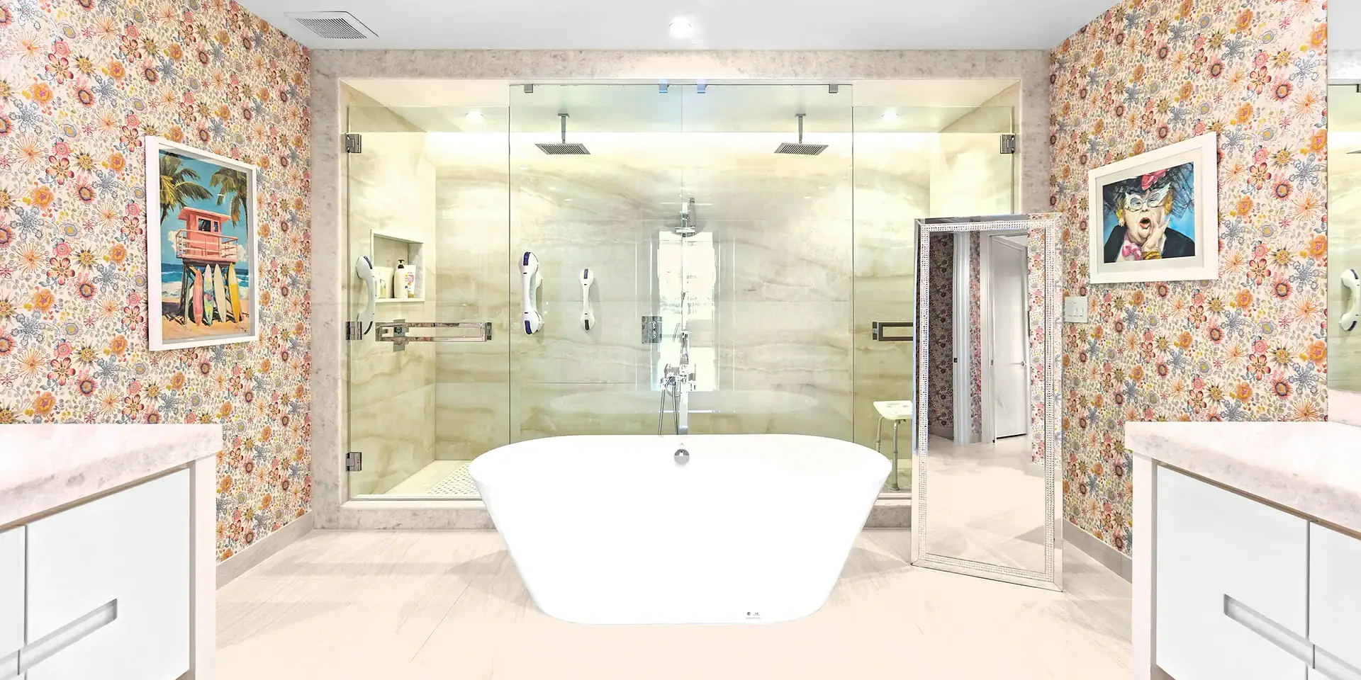 A vibrant bathroom with floral wallpaper, a white freestanding tub, a glass walk-in shower, and two framed artworks. A decorative floor mirror leans against the right wall.