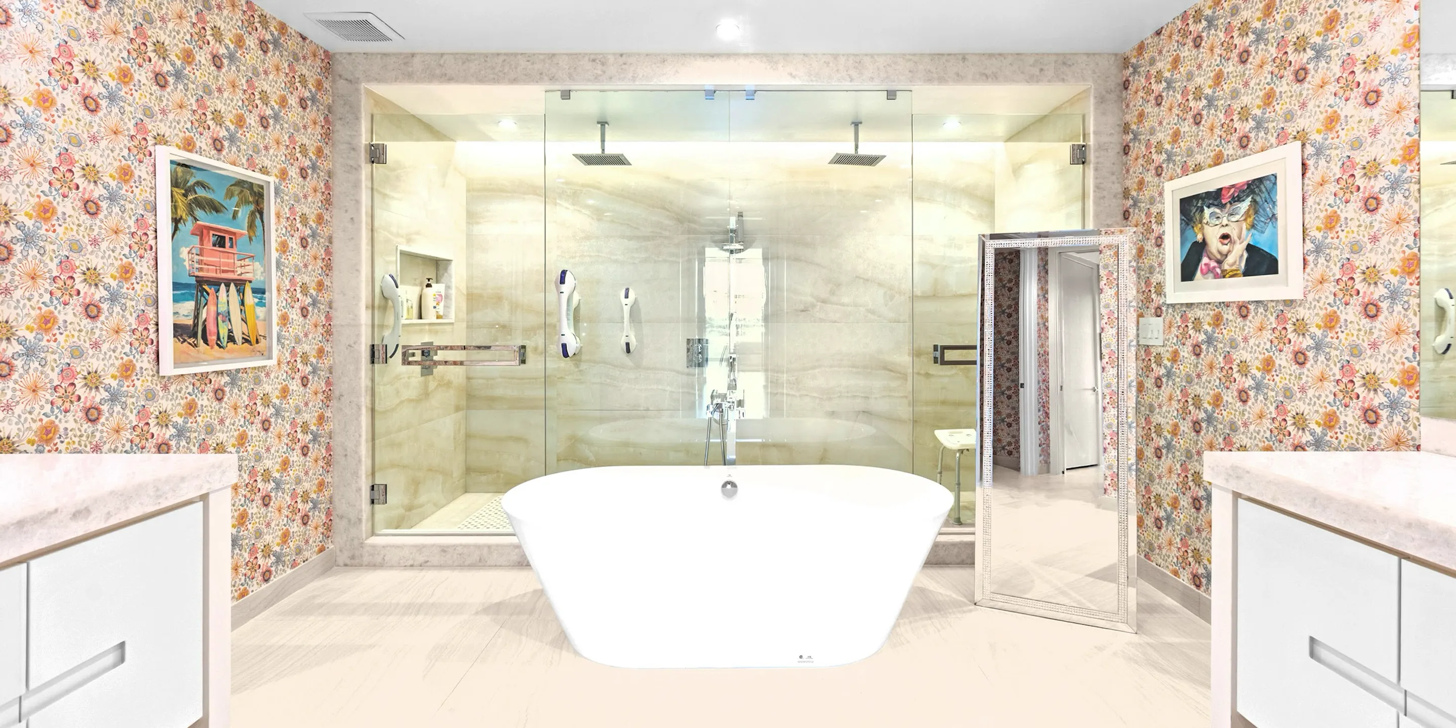 A vibrant bathroom with floral wallpaper, a white freestanding tub, a glass walk-in shower, and two framed artworks. A decorative floor mirror leans against the right wall.
