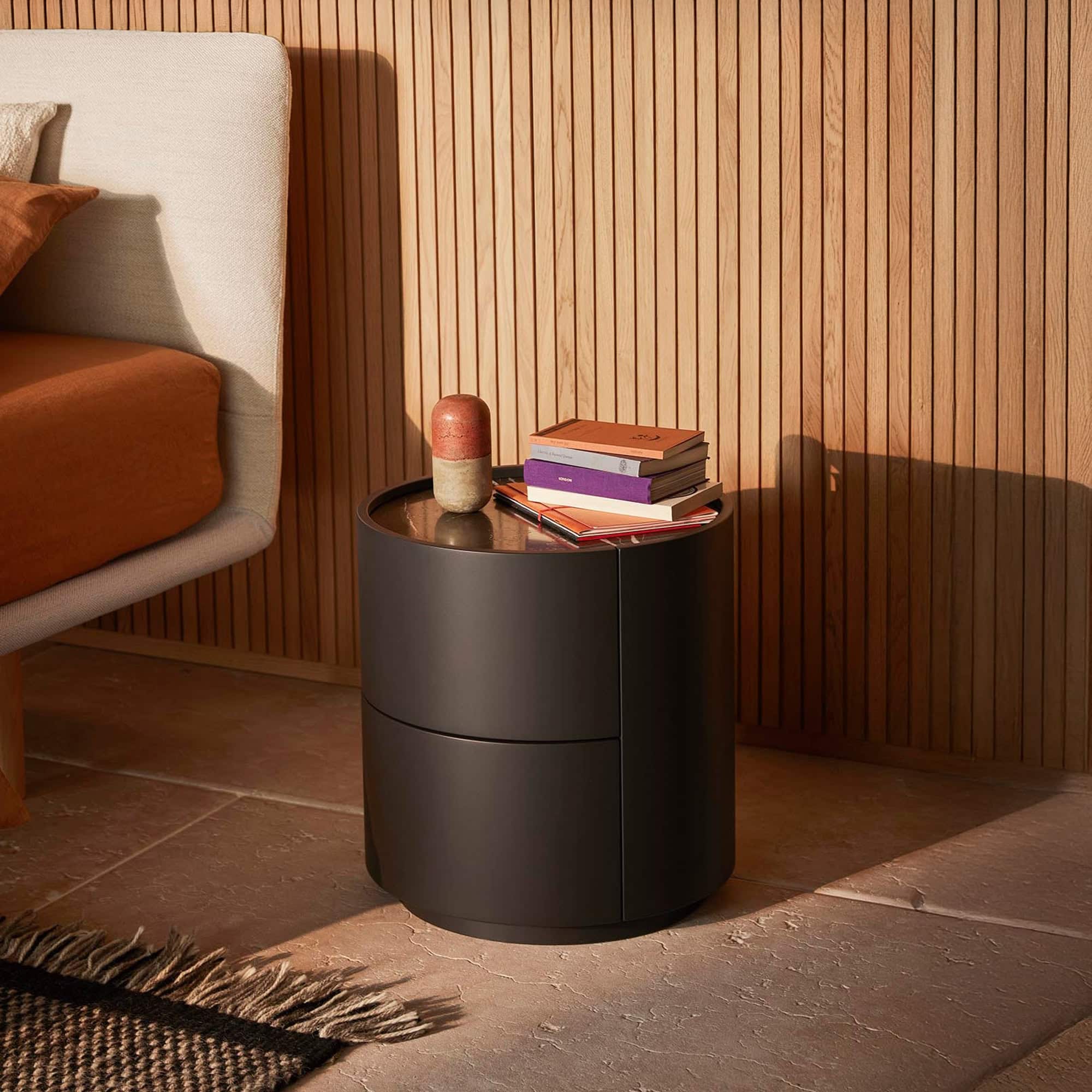 A modern bedroom scene with a dark gray cylindrical bedside table, a bed with terracotta bedding, and a wooden slatted wall, all illuminated by natural sunlight.