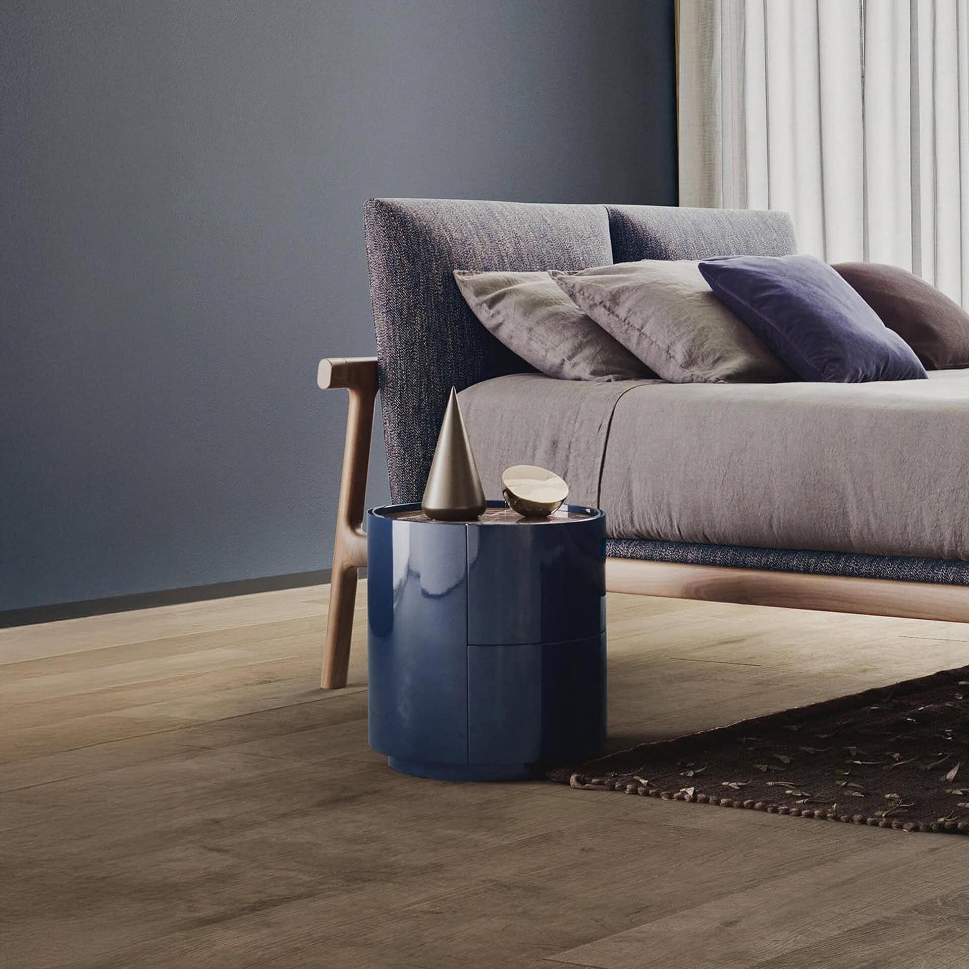 A modern bedroom with a gray-blue upholstered bed, light gray bedding, and a deep blue cylindrical nightstand with metallic decor. The wall is dark blue-gray, and the floor is light wood with a dark rug.