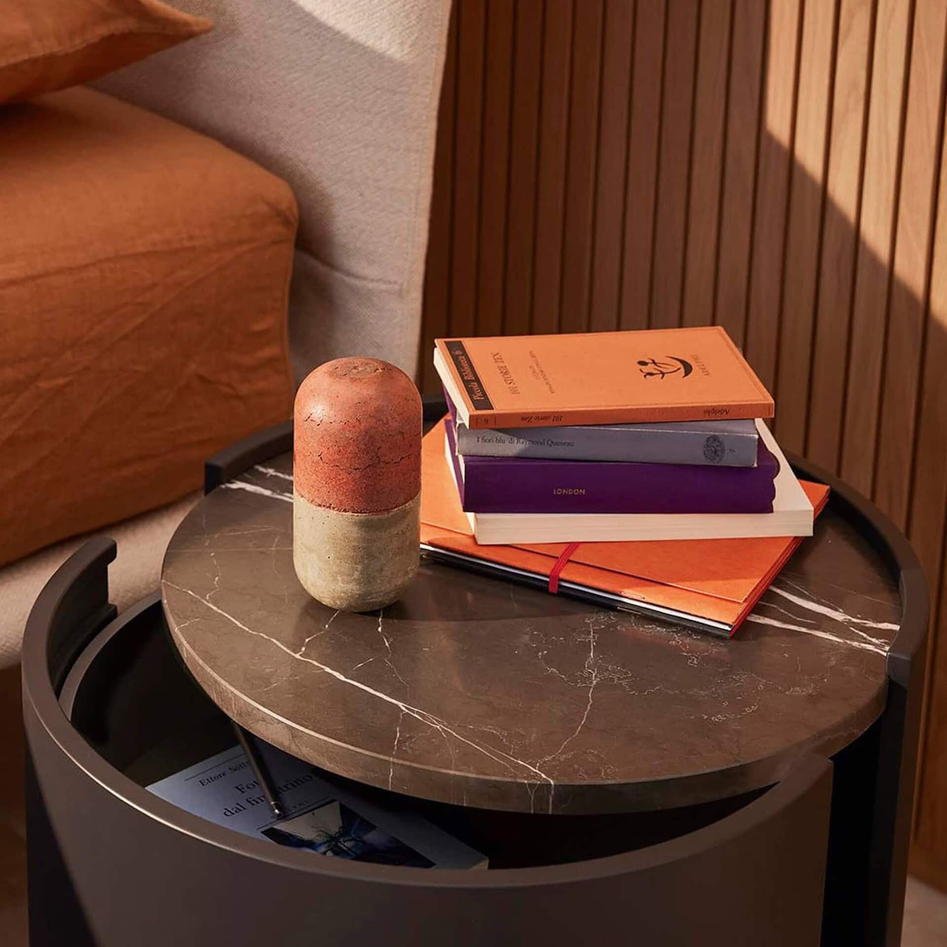 A modern dark marble side table with a two-toned capsule-shaped decor piece and a stack of books, including '101 STORIE ZEN' and 'I fiori blu di Raymond Queneau', next to a rust-colored bed and wooden slatted wall.