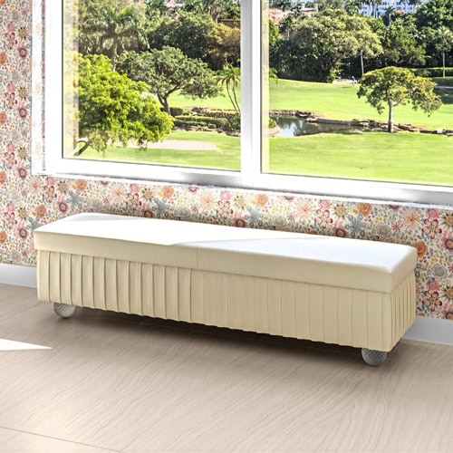 A cream-colored, pleated storage ottoman sits in front of a large window overlooking a green golf course with trees, a pond, and distant buildings. The wall has floral wallpaper.