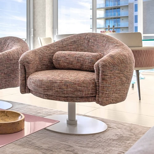 Modern swivel armchair upholstered in vibrant multi-colored tweed fabric, featuring a curved back and white circular base, set on a patterned rug with pink and white bar stools in the background.