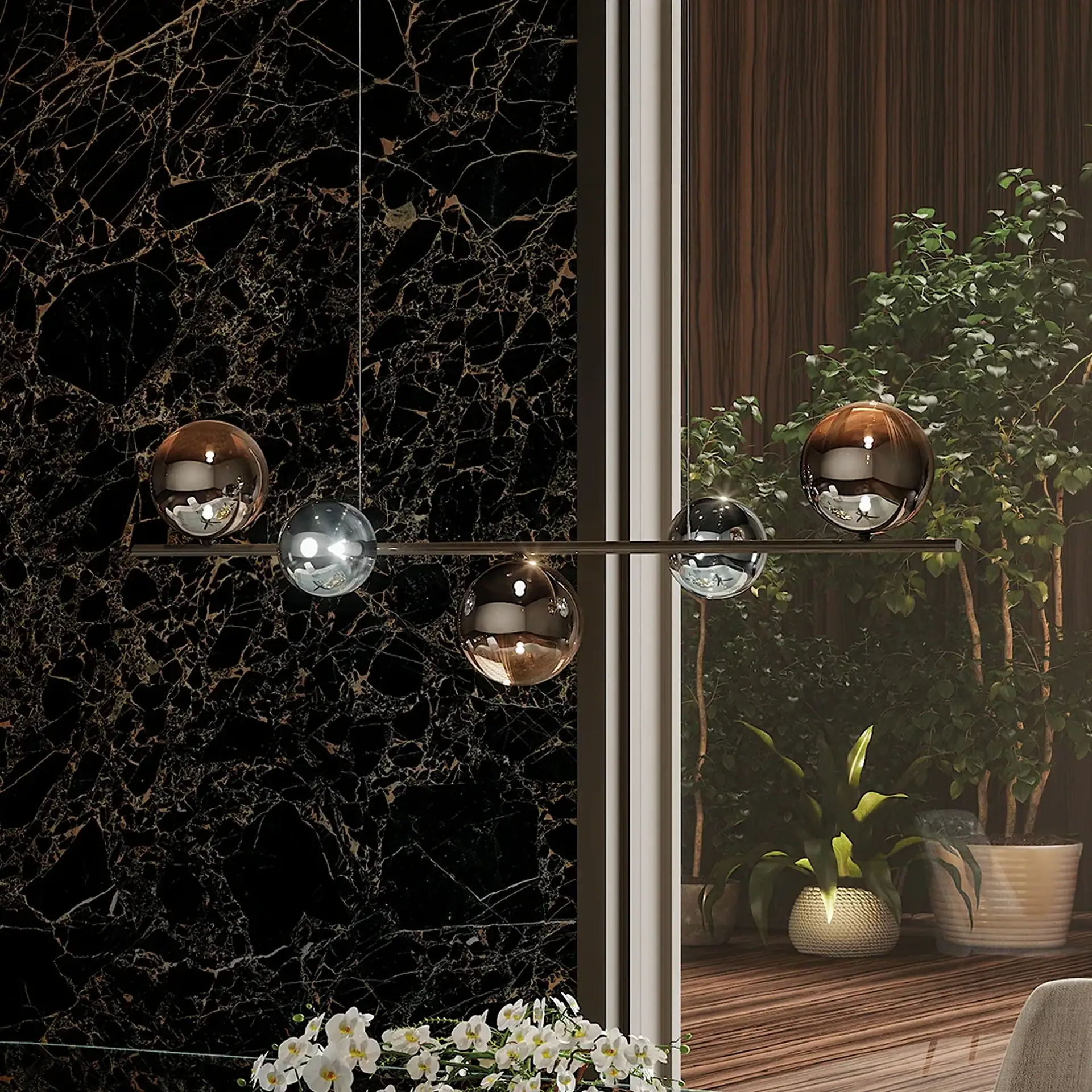 A modern linear pendant light with five spherical shades, some amber and some silver, hangs against a black marble wall with gold veins and a window overlooking green plants.