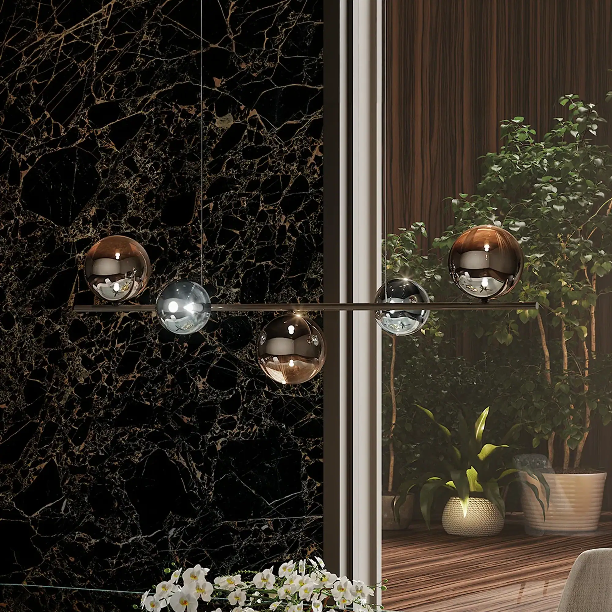 A modern linear pendant light with five spherical shades, some amber and some silver, hangs against a black marble wall with gold veins and a window overlooking green plants.