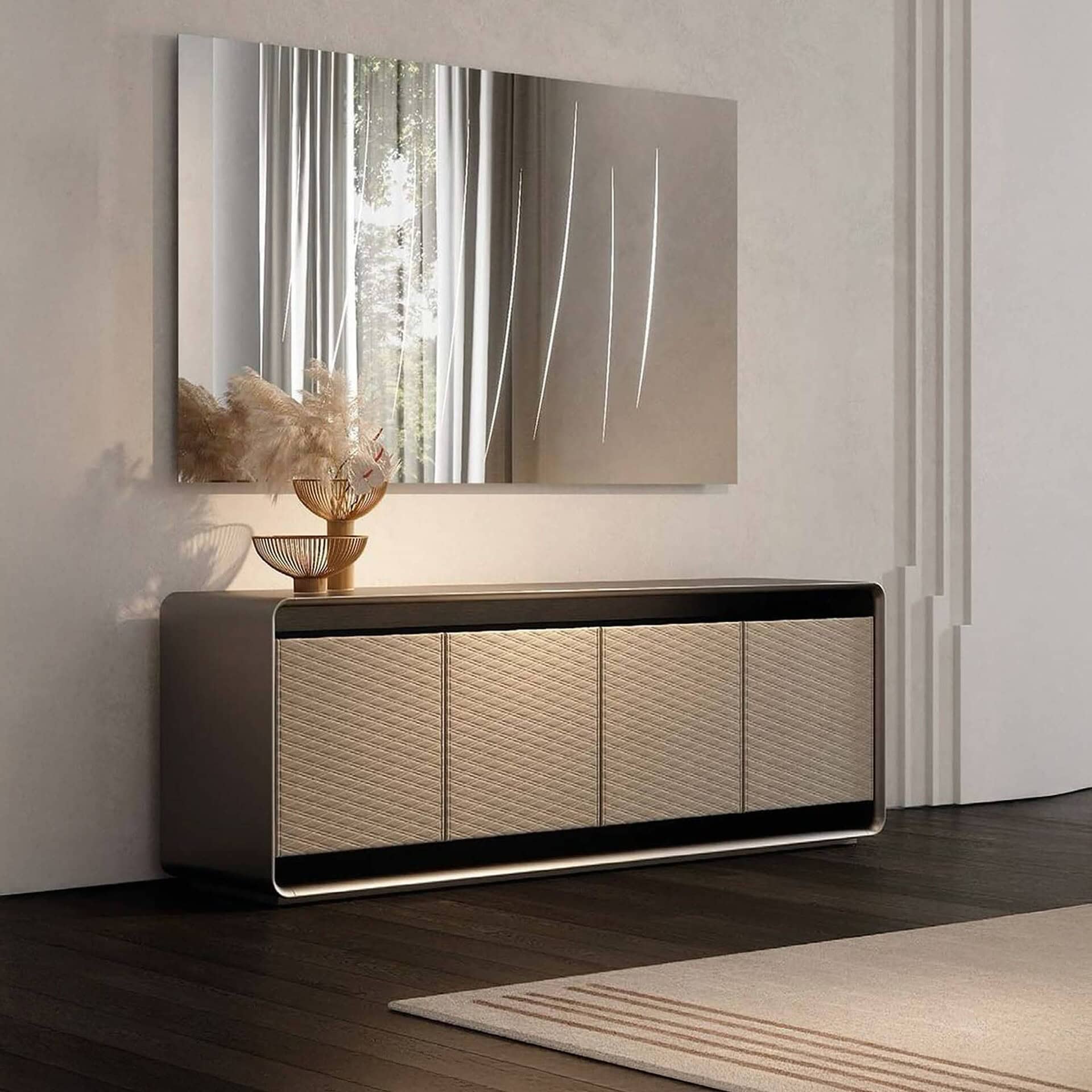 A modern living space featuring a long, dark credenza with light, textured woven doors. Above it, a large rectangular mirror reflects a window with greenery and displays several thin, vertical light streaks. On the credenza, a golden two-tiered vase holds fluffy pampas grass, illuminated by a warm light from beneath the mirror. Dark wooden floors and a light rug complete the refined look.