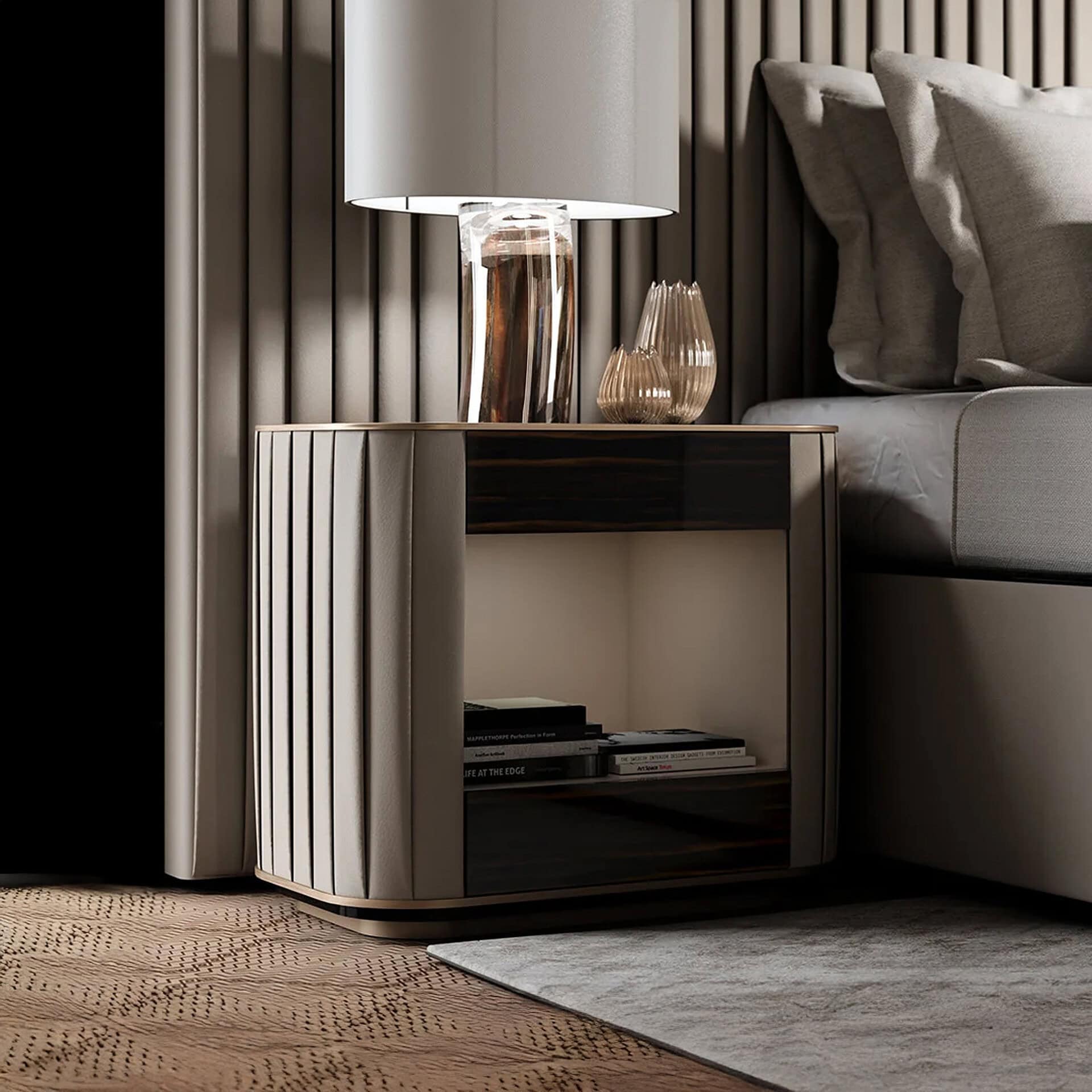 A modern bedroom scene featuring a pleated beige bedside table with a lamp, two amber vases, and books, positioned next to a grey upholstered bed and a fluted wall.