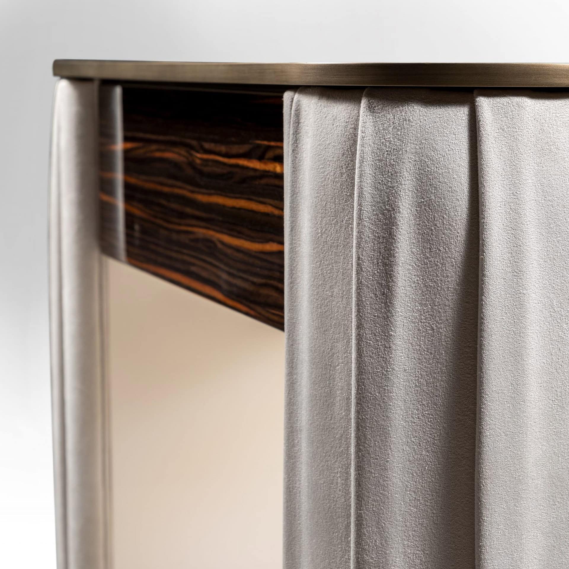 Close-up of a furniture detail featuring dark wood with prominent orange grain, light gray draped fabric, and a brushed bronze metal trim.
