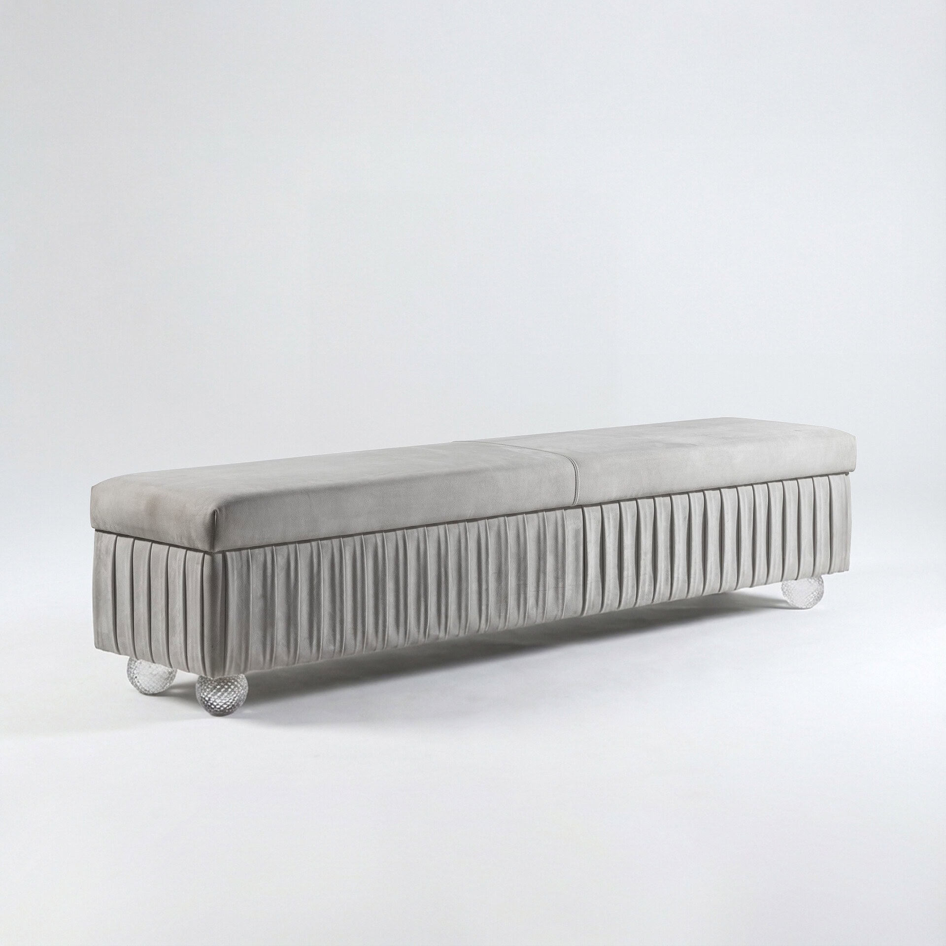 A long, light grey velvet bench with a pleated skirt design and clear, textured spherical legs, set against a plain light background.