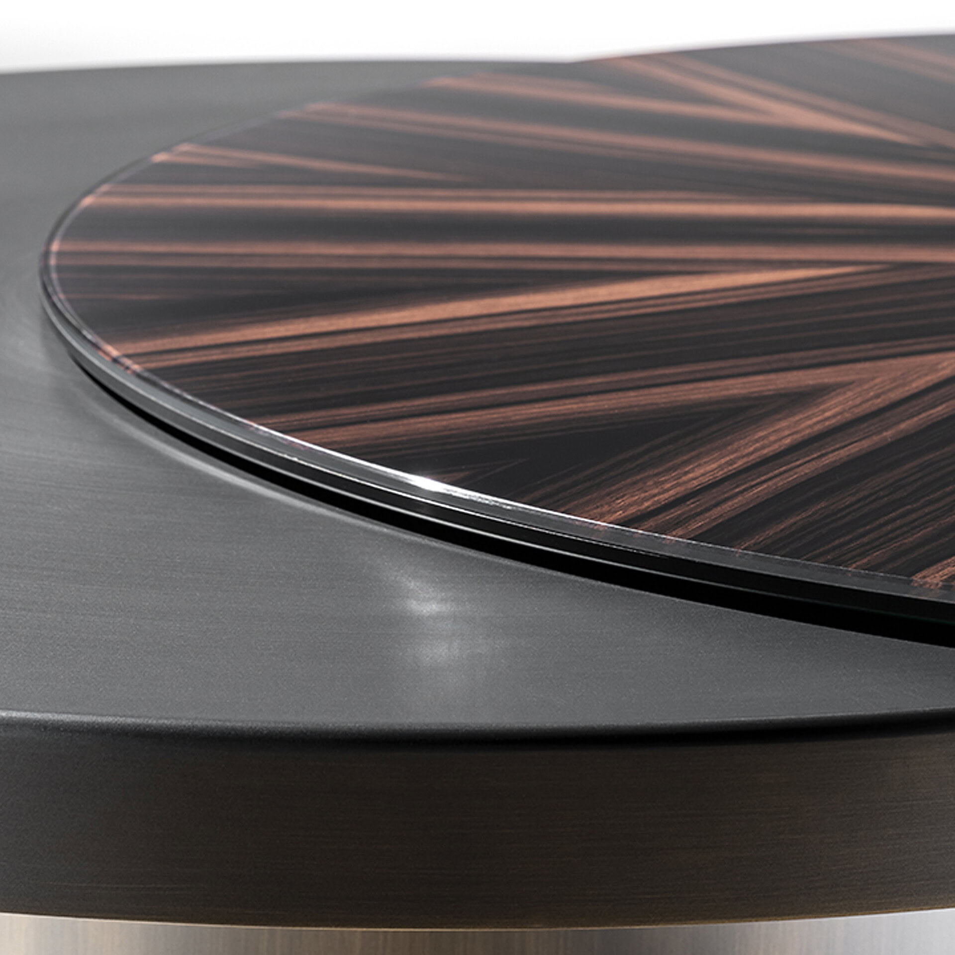 Close-up of a dark, brushed metal tabletop with a circular sunburst wood veneer insert featuring radiating dark and reddish-brown lines, separated by a thin metallic trim.