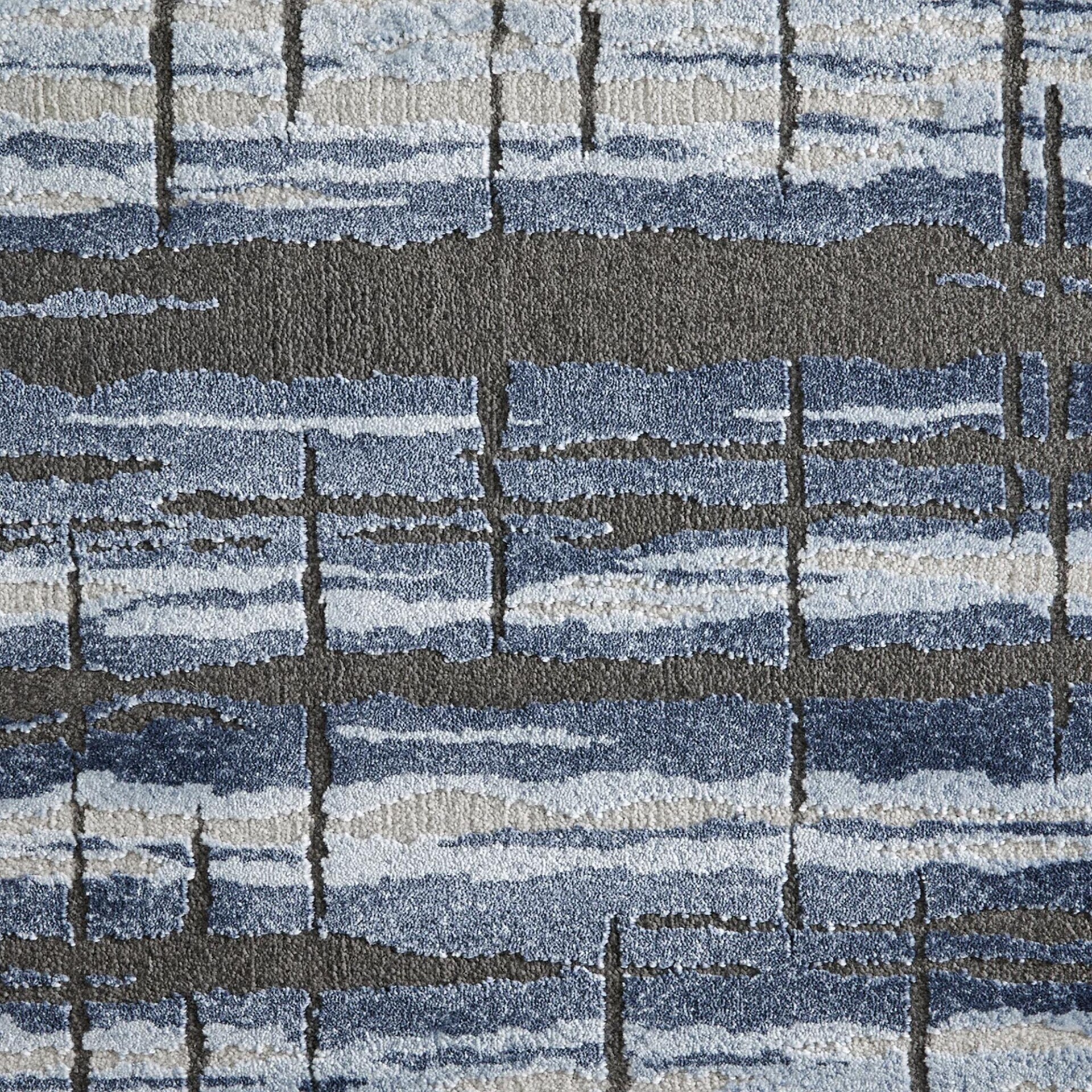 Close-up of a textured carpet with an abstract pattern of irregular horizontal bands in shades of blue and beige, intersected by broken vertical lines of dark gray.