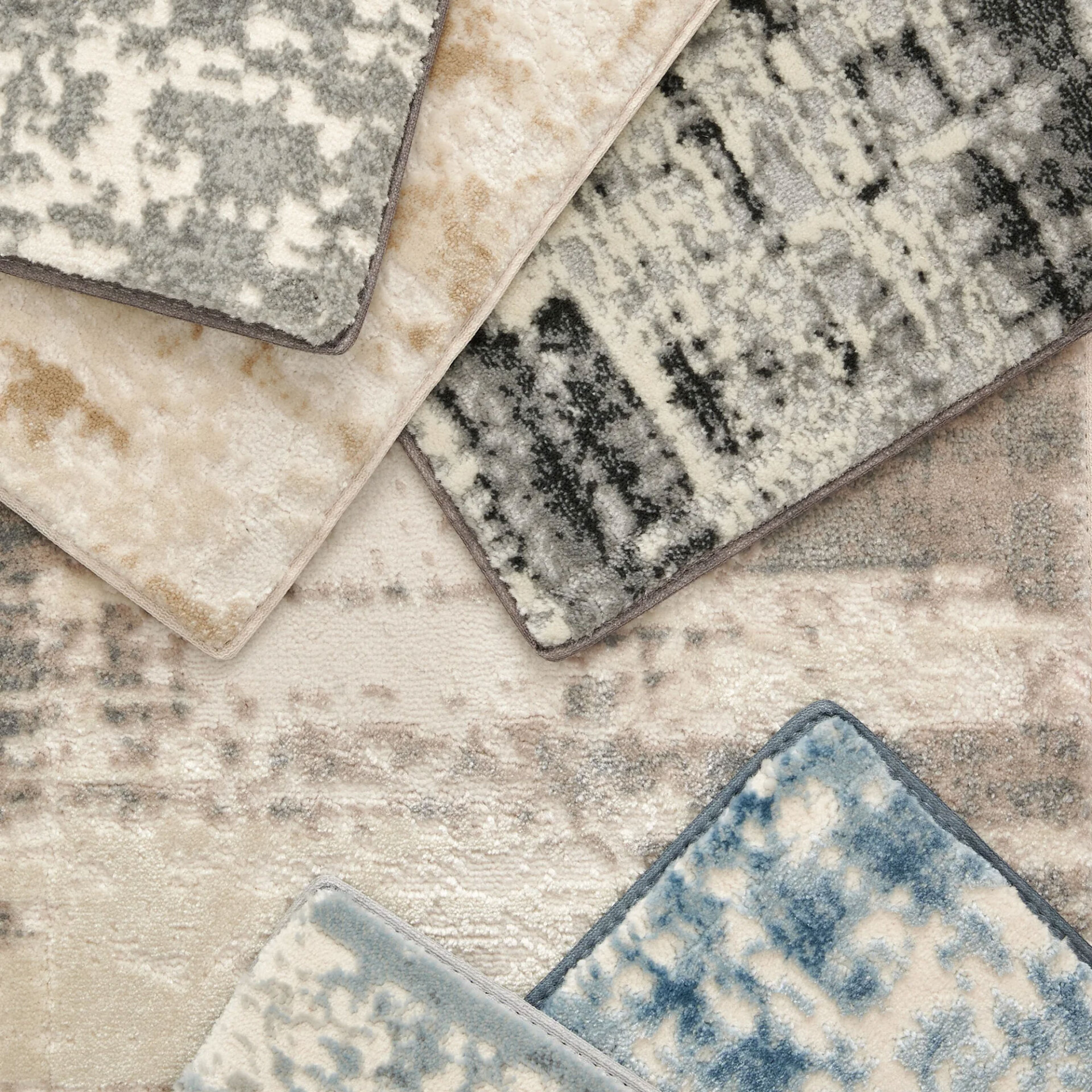 A flat lay of overlapping rug samples in various abstract patterns and textures. Colors include white, cream, beige, grey, black, and two shades of blue.