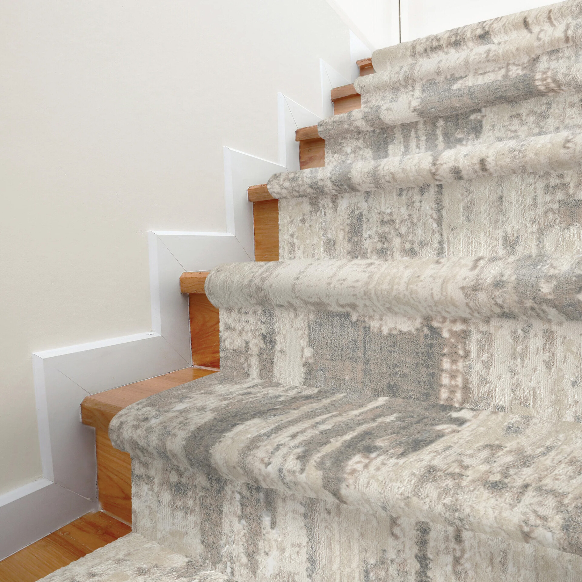 A modern staircase featuring light wooden treads and a cream, beige, and grey abstract patterned carpet runner. White trim lines the wall.