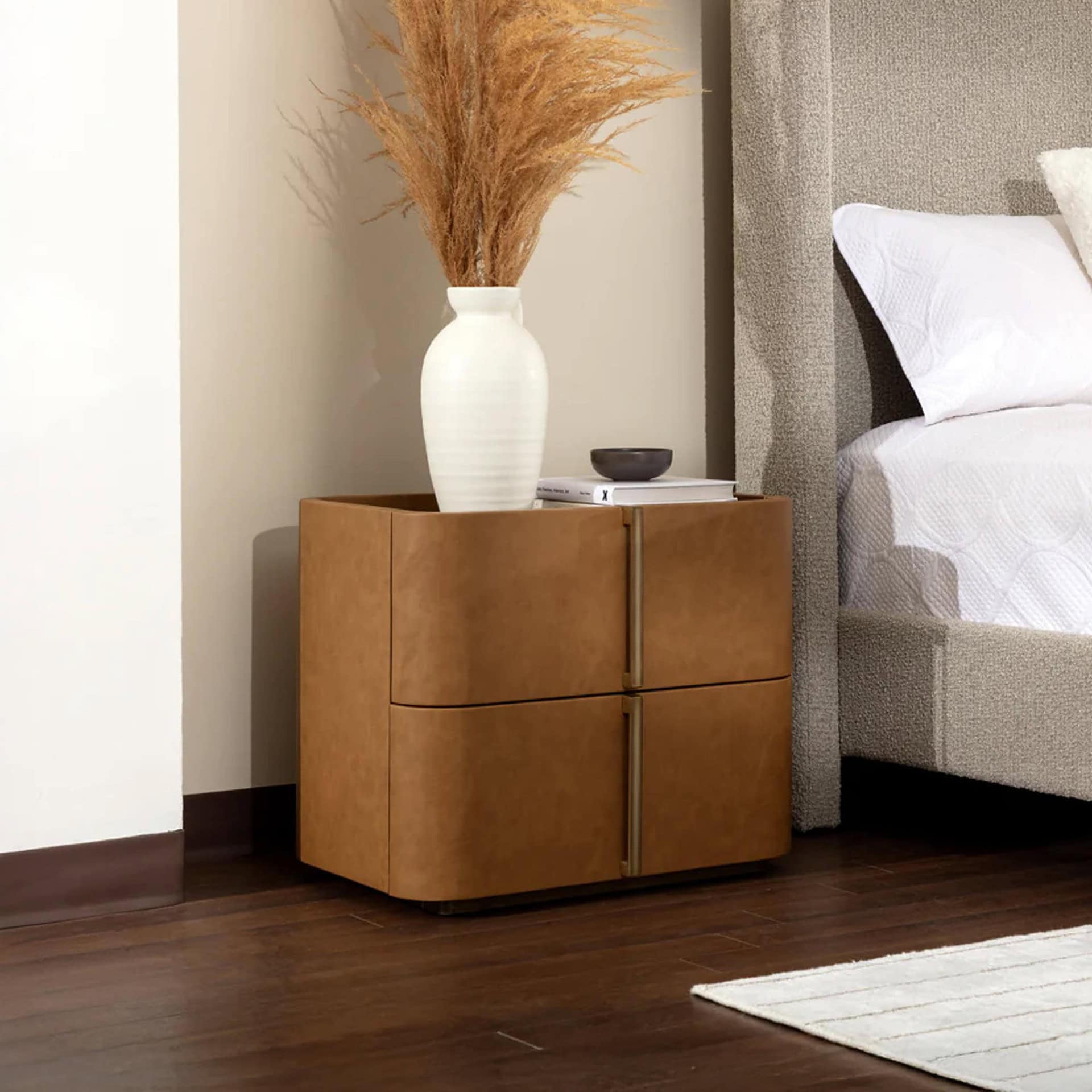 A modern bedroom scene featuring a tan leather bedside table with pampas grass in a white vase, next to a bed with a grey upholstered headboard and white bedding. Dark wood floors are visible.