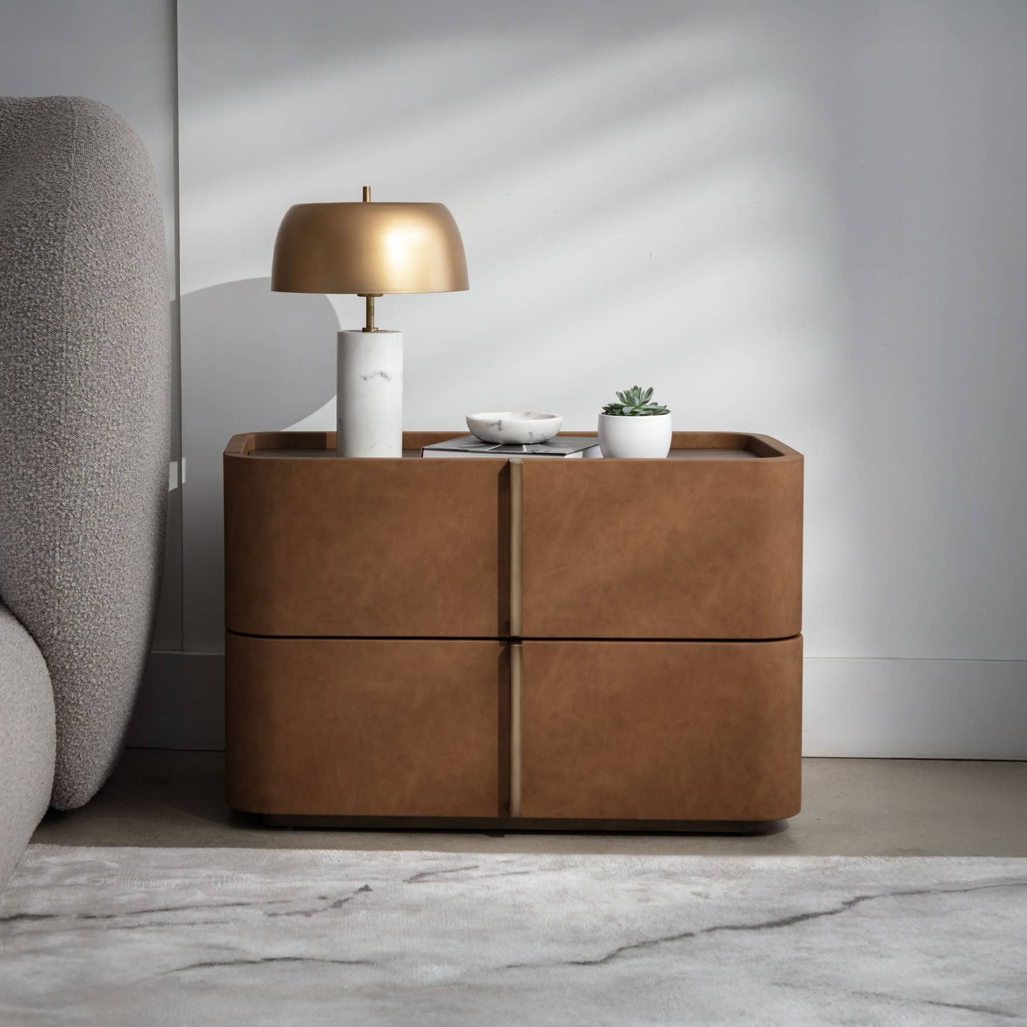 A modern brown leather nightstand with two drawers, topped with a gold and marble lamp, a succulent, and a marble bowl on books. A gray textured headboard is to the left, and a white and gray rug is on the floor.