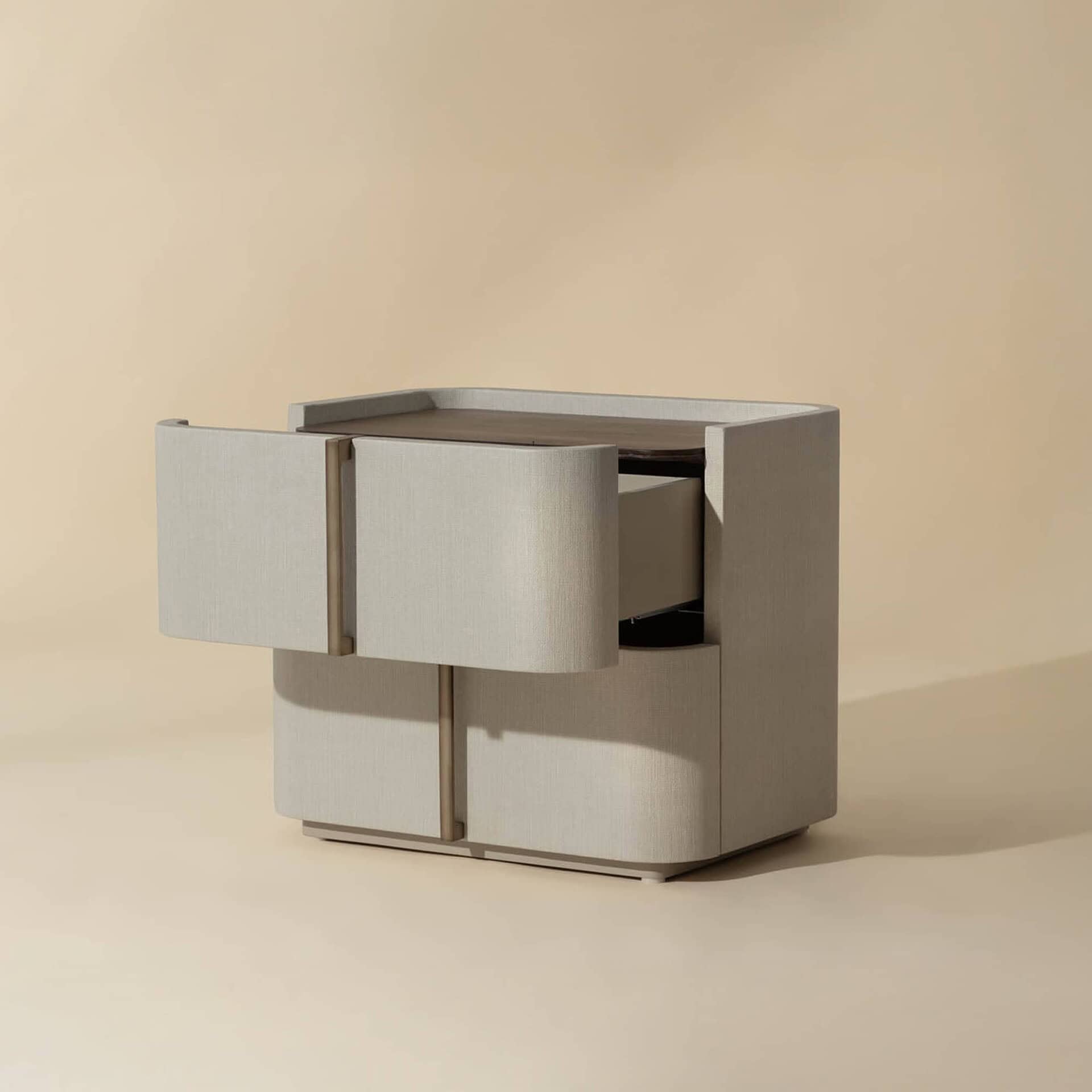 A modern nightstand with a light textured fabric finish, curved front, and brass handles. The top drawer is partially open, revealing its interior.