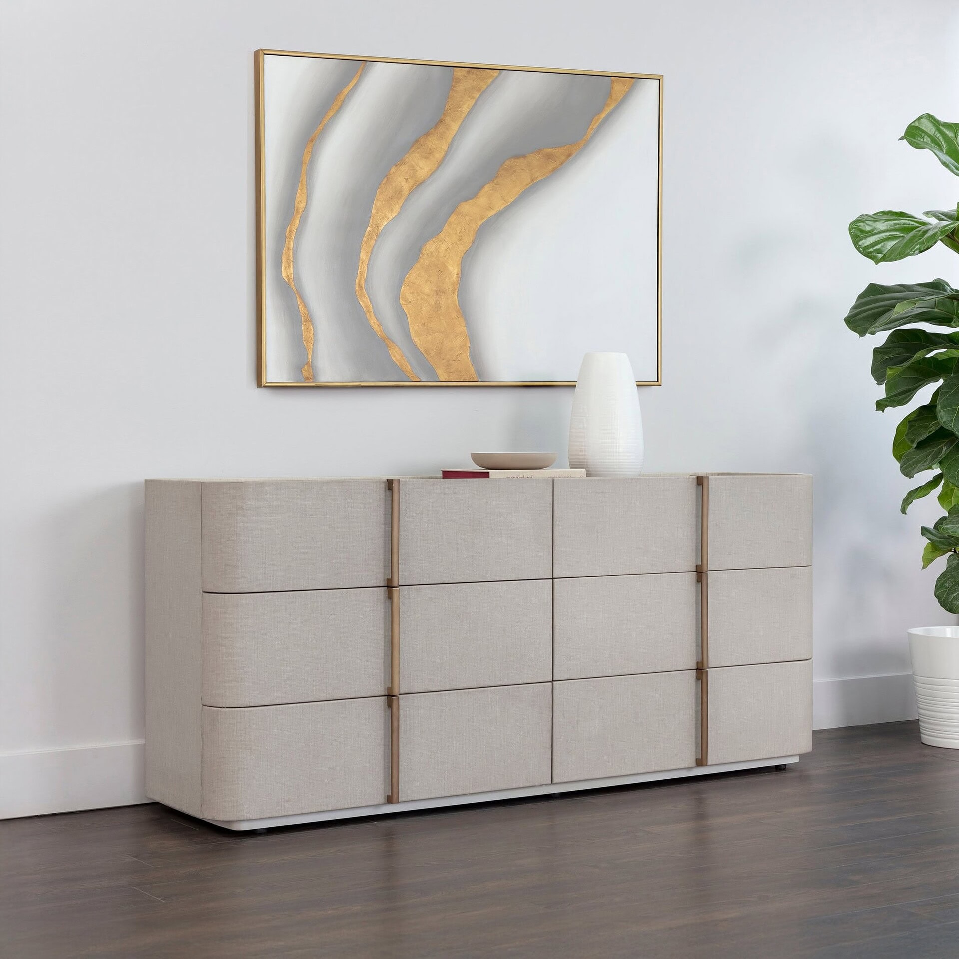A modern beige upholstered dresser with gold handles, topped with a white vase and books, sits below an abstract gold and gray painting. A fiddle leaf fig plant in a white pot is to the right, all against a white wall and dark wood floor.