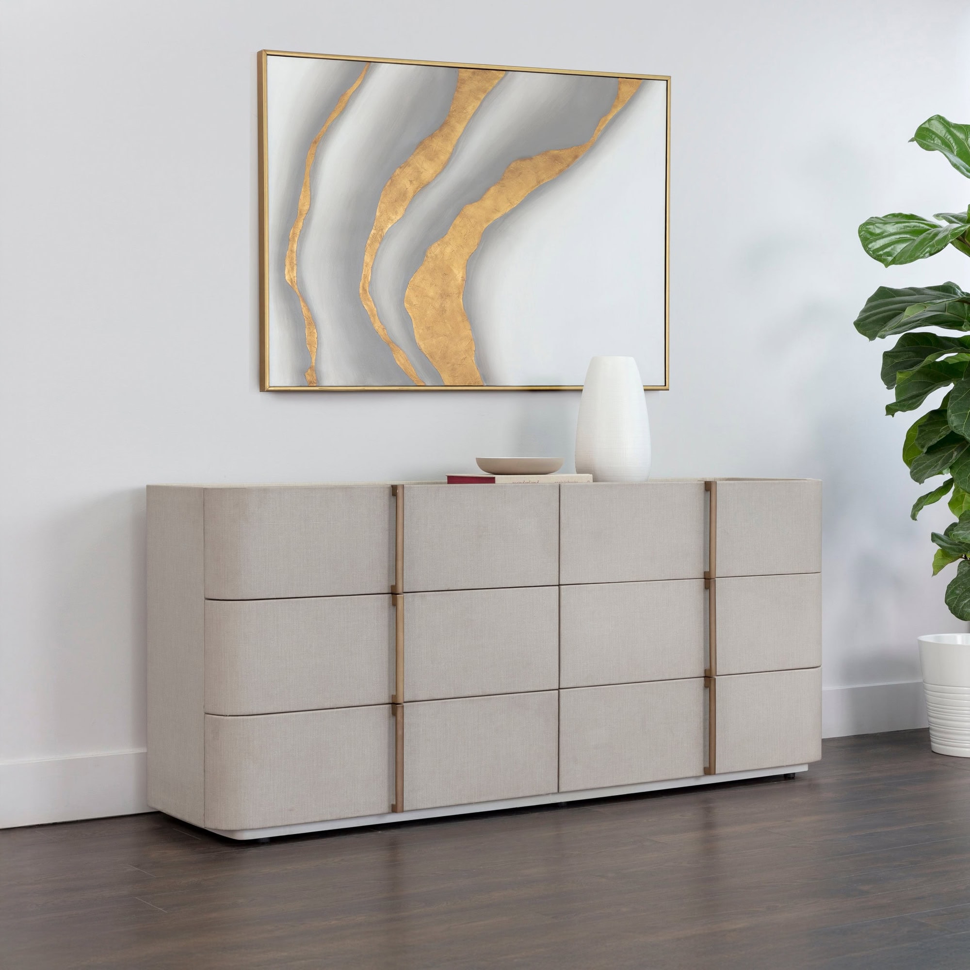 A modern beige upholstered dresser with gold handles, topped with a white vase and books, sits below an abstract gold and gray painting. A fiddle leaf fig plant in a white pot is to the right, all against a white wall and dark wood floor.