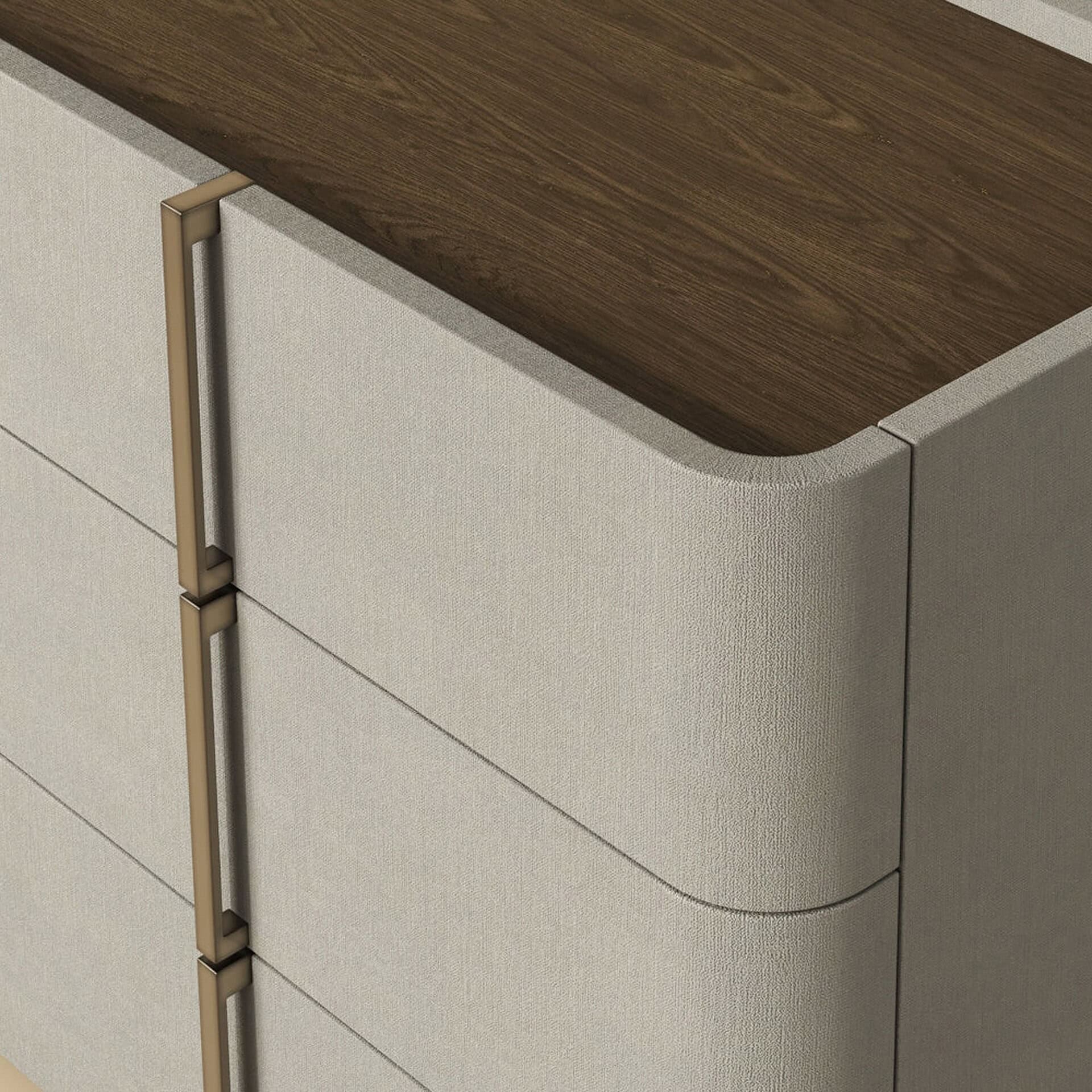 Close-up of a modern dresser with a dark wood top, light beige fabric-wrapped body with rounded corners, and vertical bronze handles.