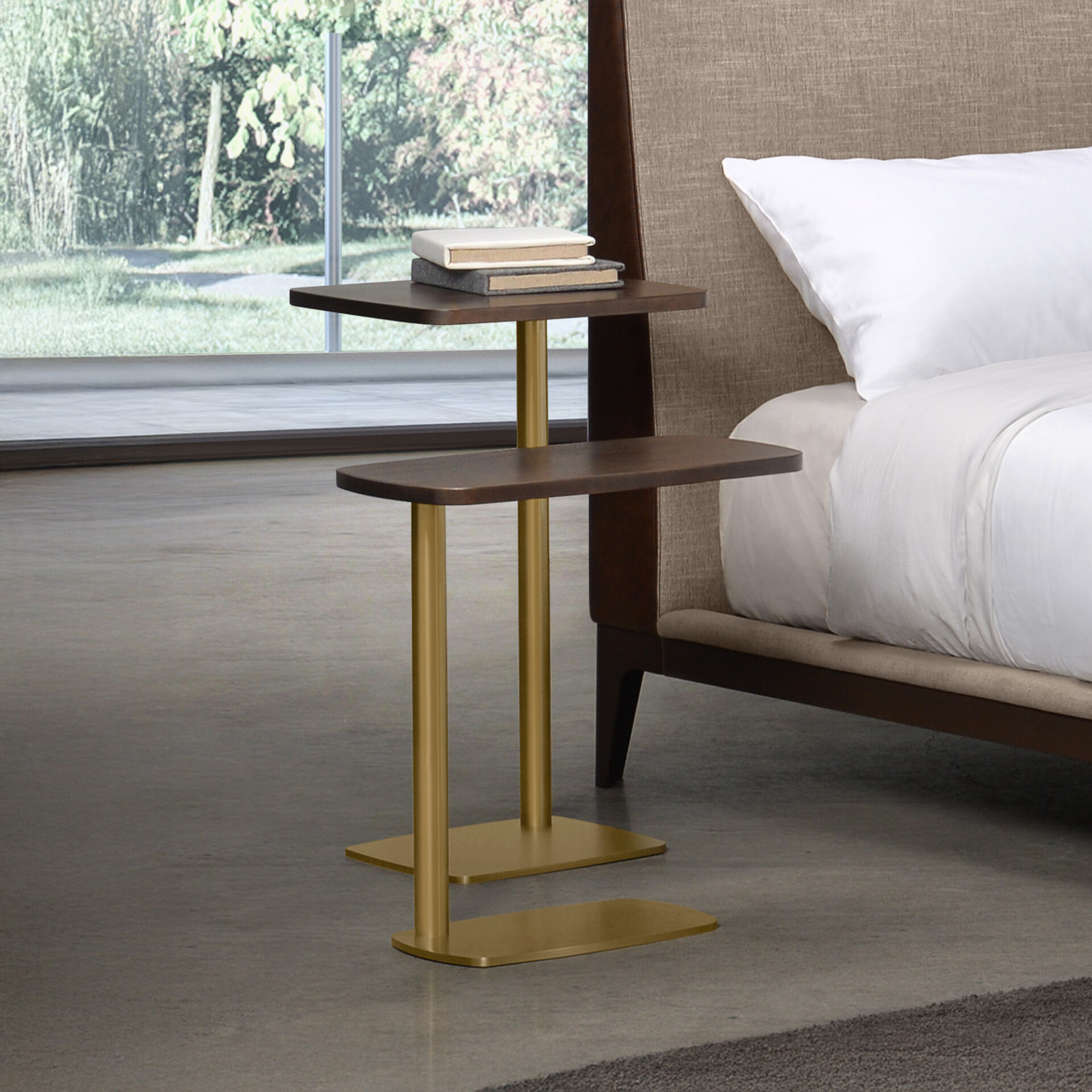 A modern, two-tiered C-shaped side table with dark wood shelves and brass legs, positioned next to a bed with a fabric headboard. Two books rest on the top shelf. A large window with a green outdoor view is in the background.
