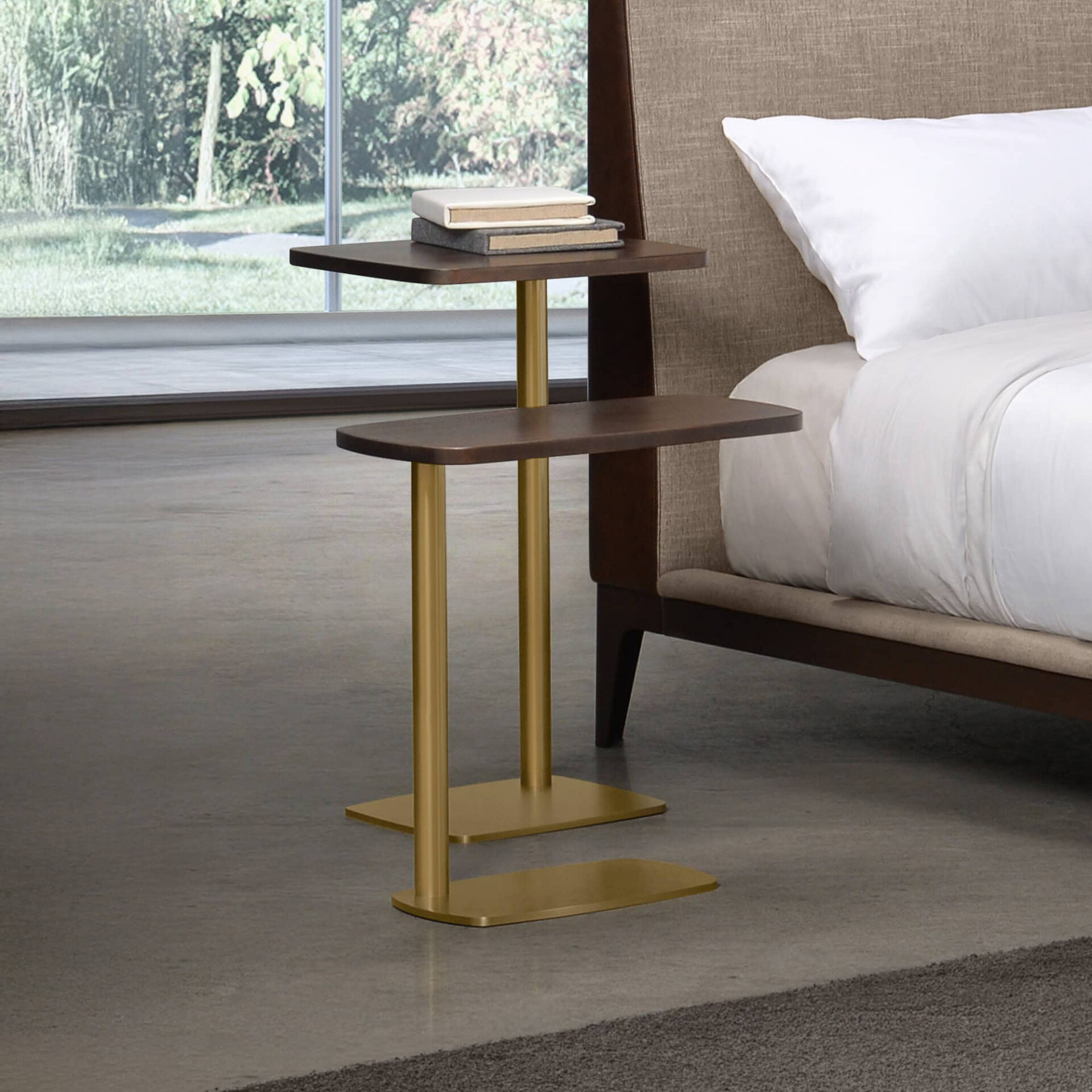 A modern, two-tiered C-shaped side table with dark wood shelves and brass legs, positioned next to a bed with a fabric headboard. Two books rest on the top shelf. A large window with a green outdoor view is in the background.