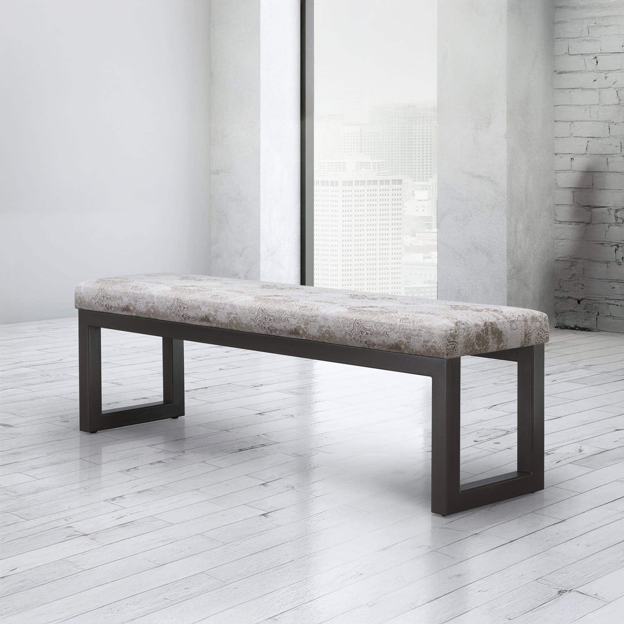 A modern upholstered bench with a patterned beige cushion and dark metal legs in a bright, minimalist room with a city view from a large window.