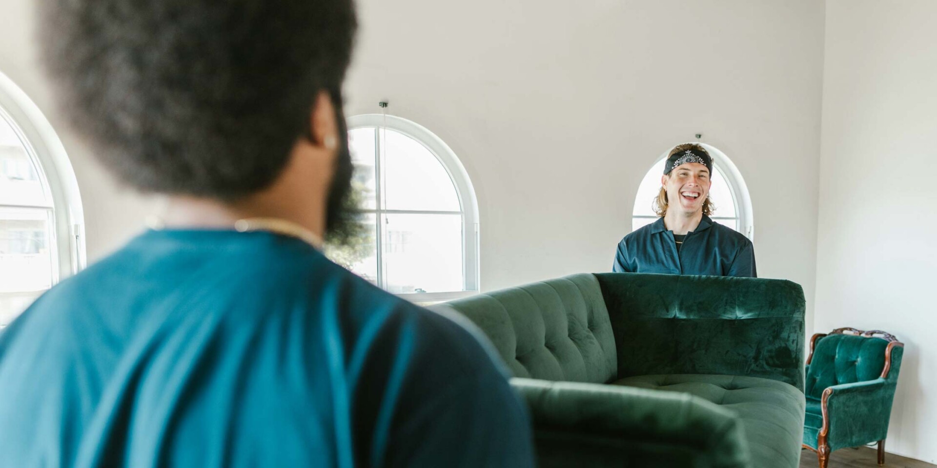 Two people moving a large, dark green velvet sofa in a bright room with arched windows. One person is seen from behind, and the other smiles broadly from behind the sofa.