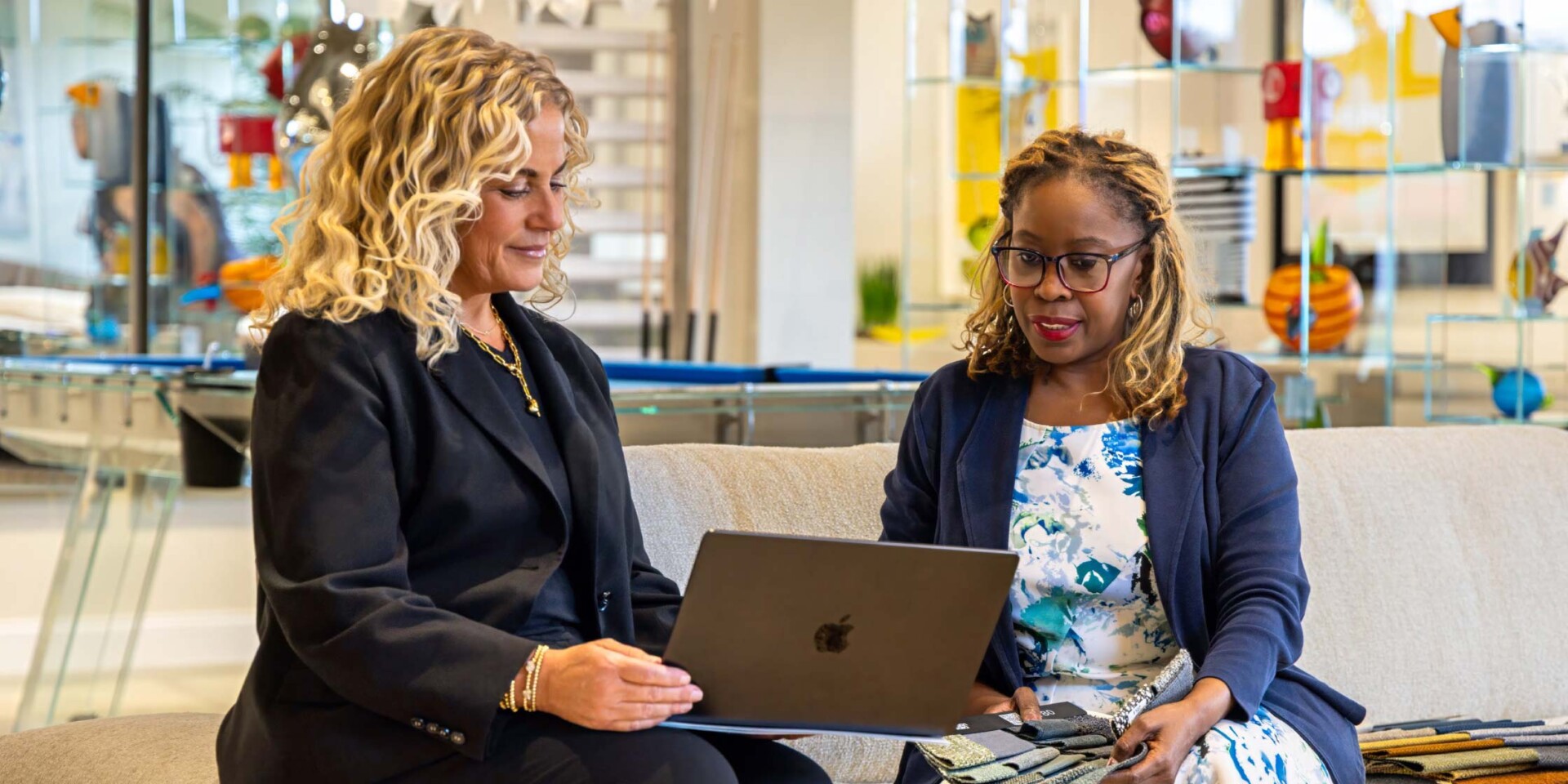 Two women sitting on a sofa, looking at a laptop. One woman holds fabric swatches, suggesting a design consultation in a modern office lounge.