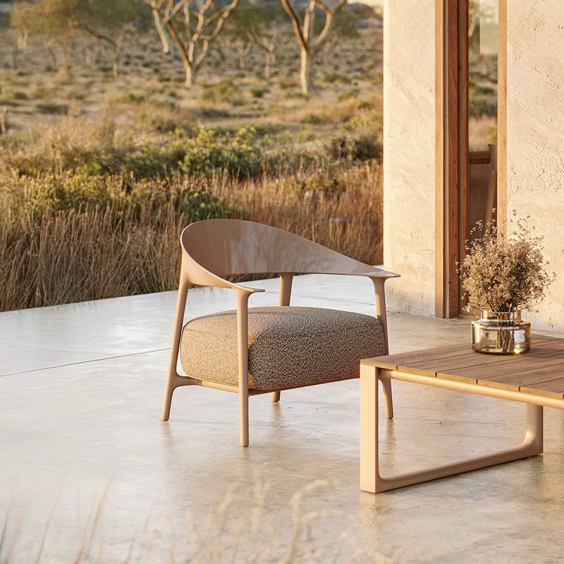 A modern outdoor patio featuring a curved wooden armchair with an upholstered seat, a slatted wooden coffee table with a vase of dried flowers, and a natural, dry landscape in the background, all bathed in warm, soft light.