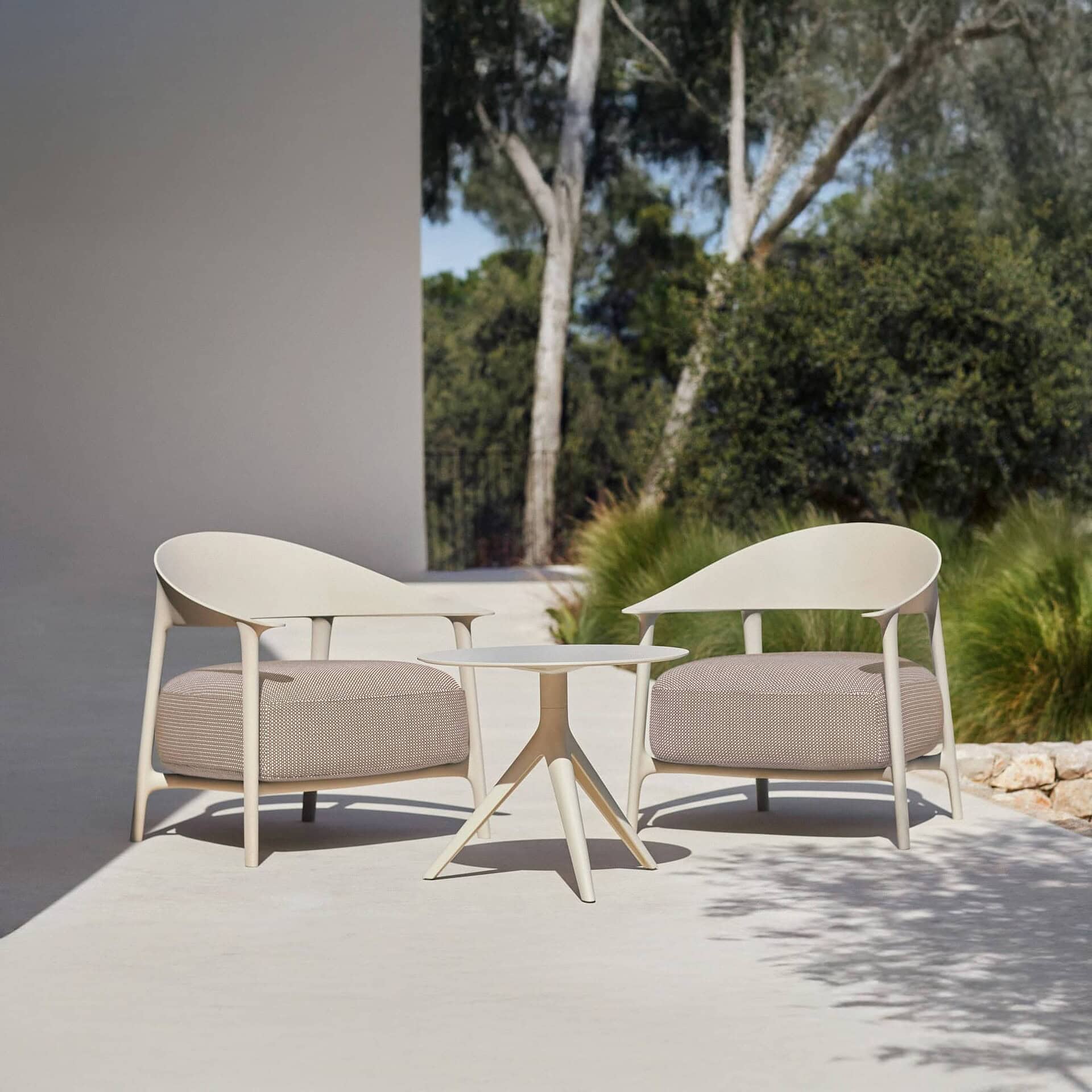 Two modern light-colored outdoor armchairs with textured cushions and a matching round side table on a patio, surrounded by lush green trees and foliage under dappled sunlight.