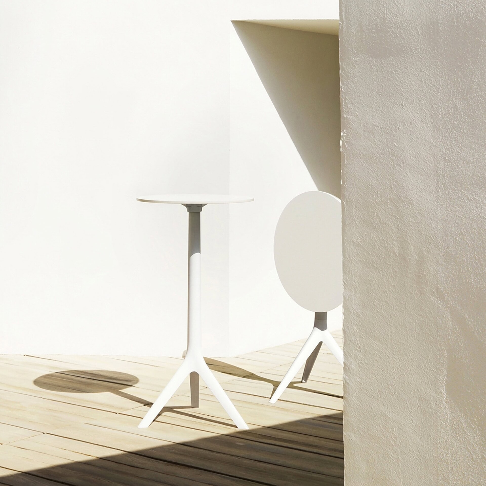 Two modern white high-top tables on a wooden deck. One table is fully visible, casting a round shadow, while the other is partially hidden behind a textured off-white wall, all under bright sunlight creating strong geometric shadows.