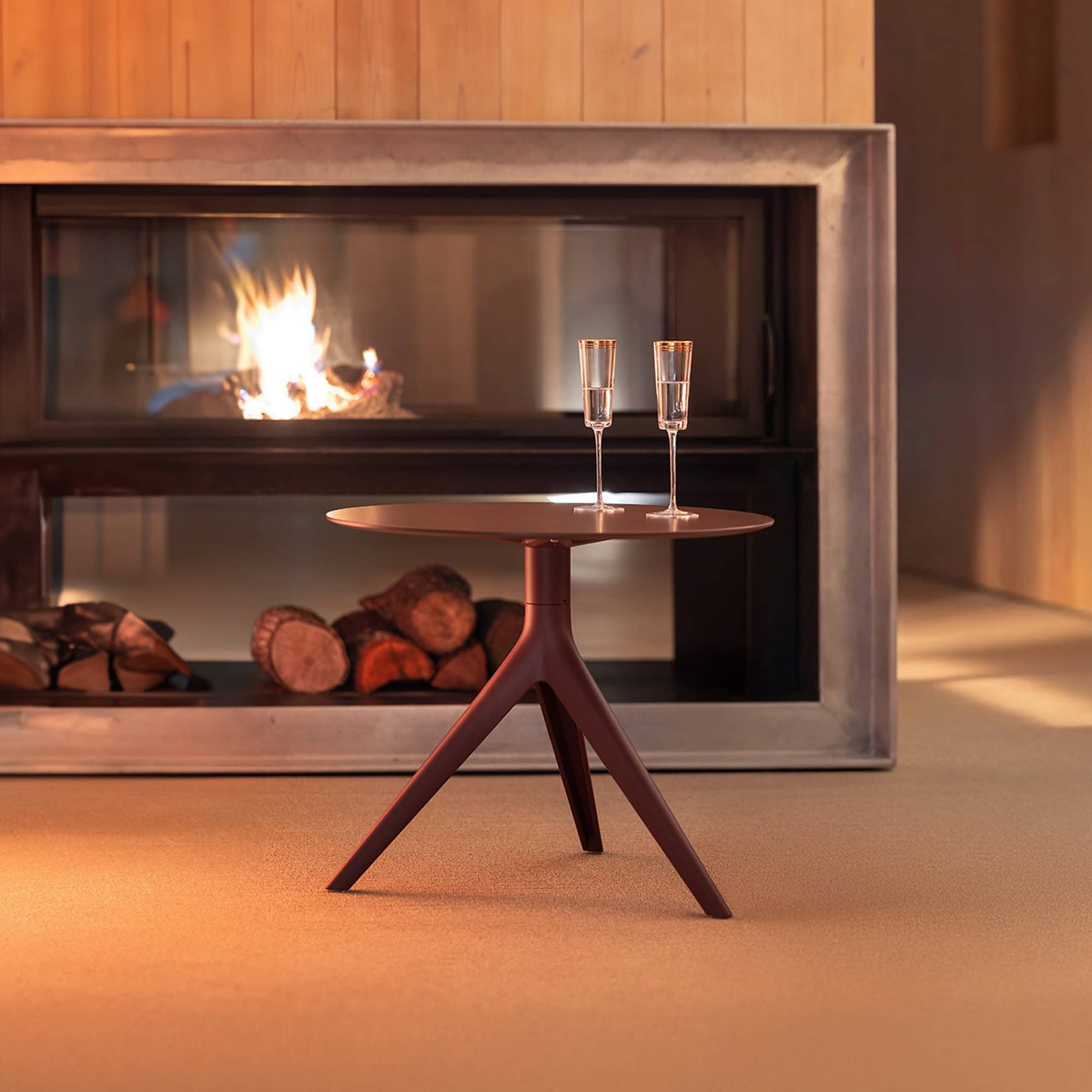 A modern, dark reddish-brown round side table with two gold-rimmed champagne flutes in front of a contemporary fireplace with a crackling fire and stacked logs.