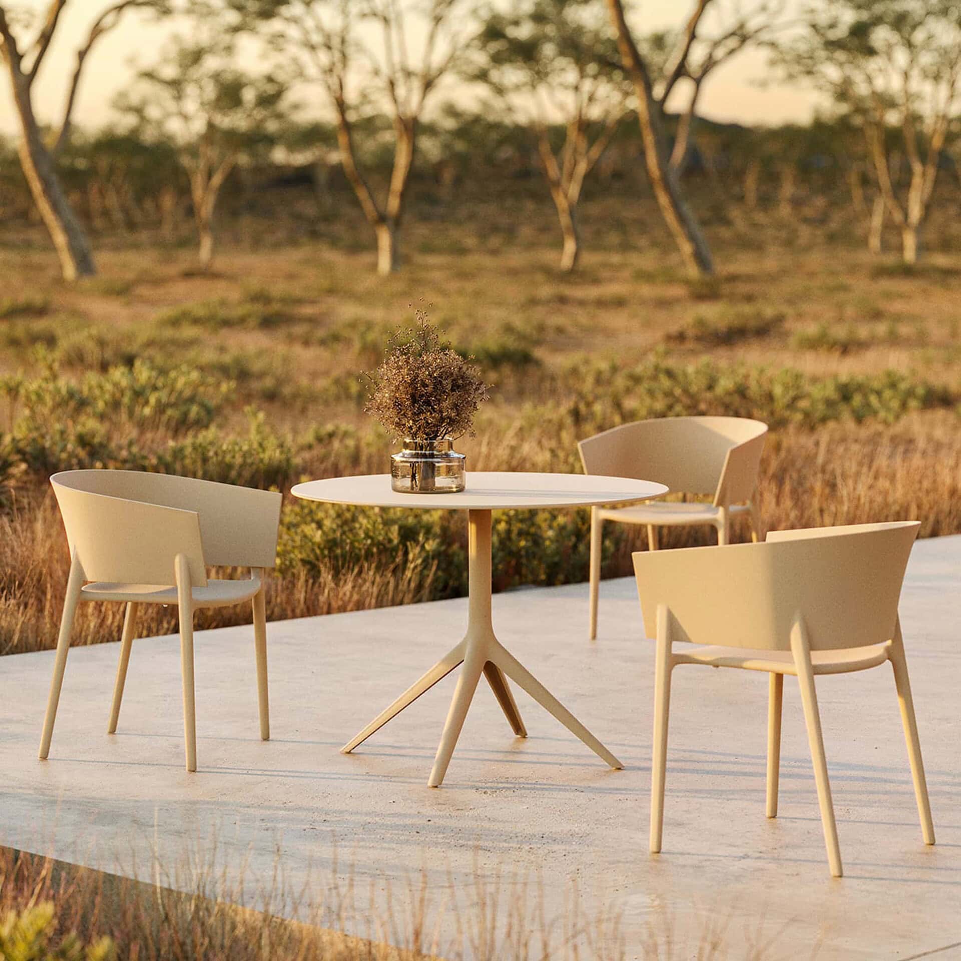 An outdoor dining set with a round light beige table and three matching chairs on a concrete patio. A vase with dried plants sits on the table. The scene is bathed in warm, golden light, with a natural, arid landscape and sparse trees in the background.