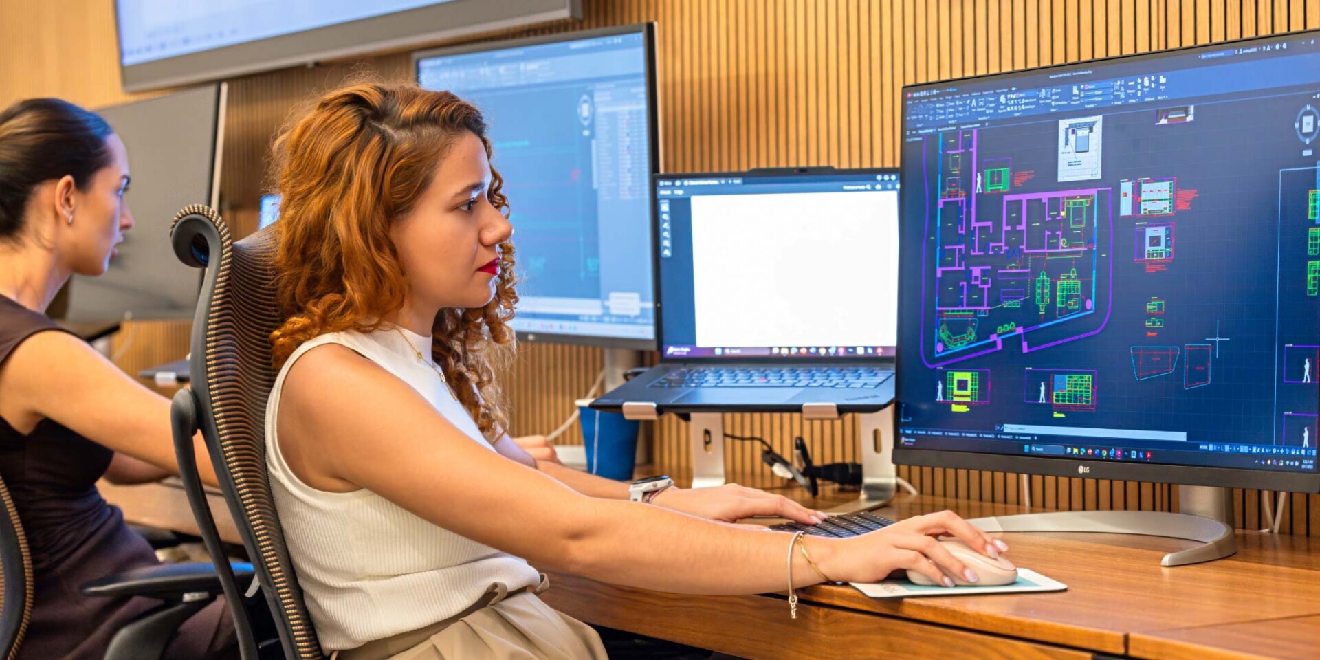Two women working at computer workstations in an office. The foreground woman with curly hair is intently using CAD software displaying architectural floor plans on a large monitor.