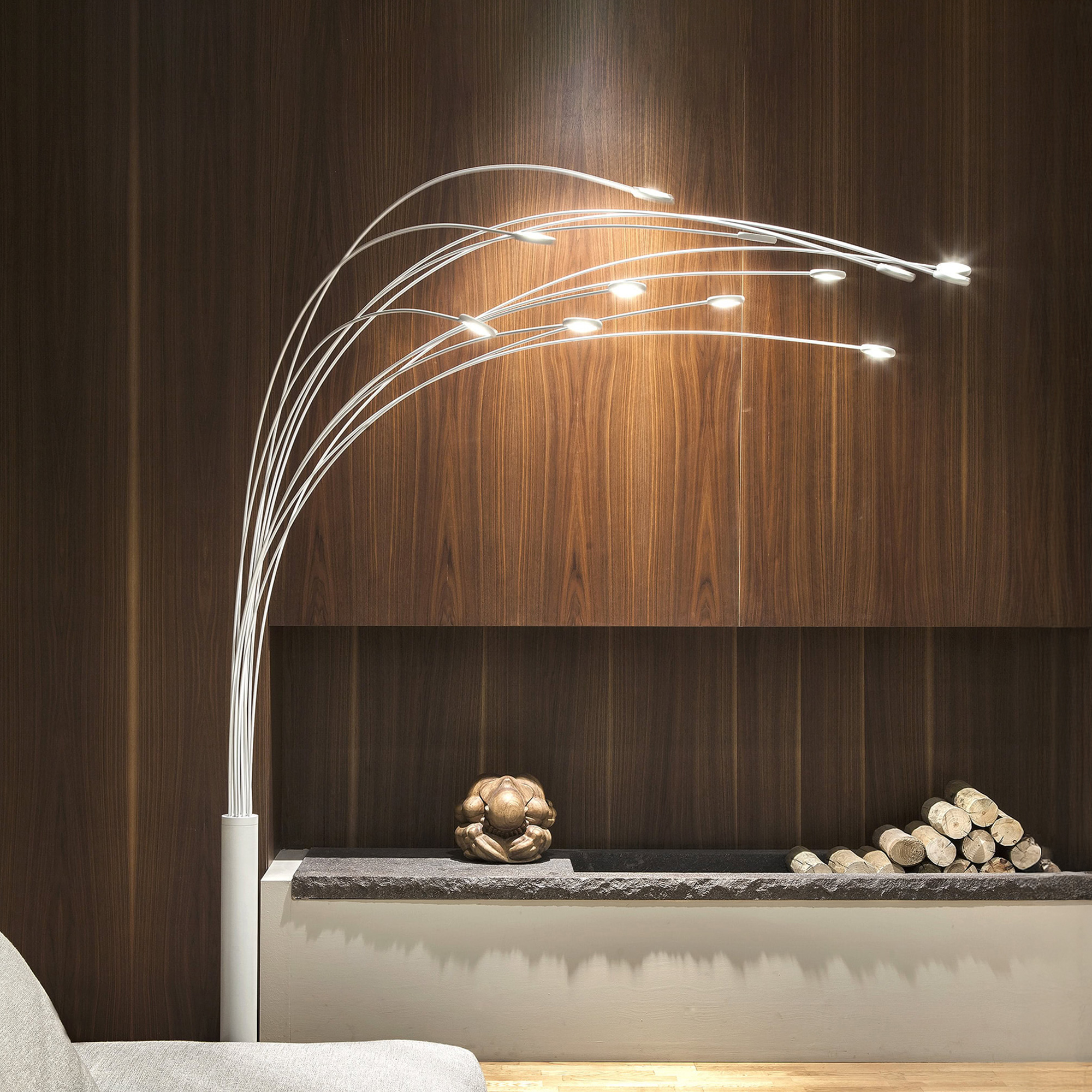 A modern, sculptural floor lamp with multiple arched arms and LED lights, standing next to a dark wooden paneled wall. Below the paneling is a low shelf with a dark stone top, holding an abstract wooden sculpture and a stack of logs.