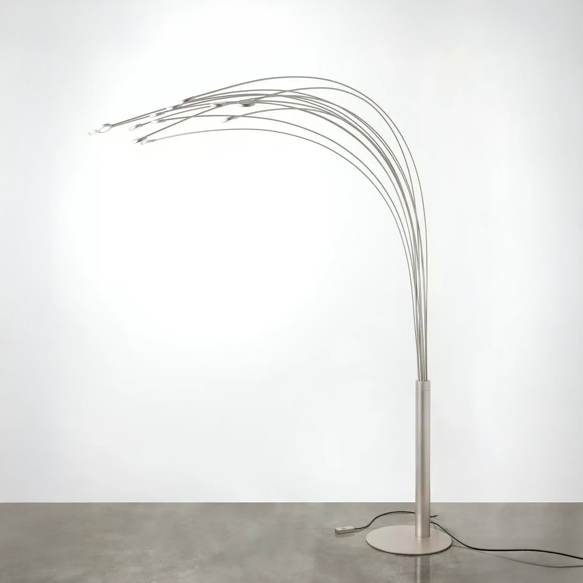 A modern, silver-toned floor lamp with a circular base, a slender stem, and multiple thin, arching metal rods that curve upwards and then downwards, each tipped with a small, illuminated LED light. The lamp is set against a plain white wall and a grey floor.