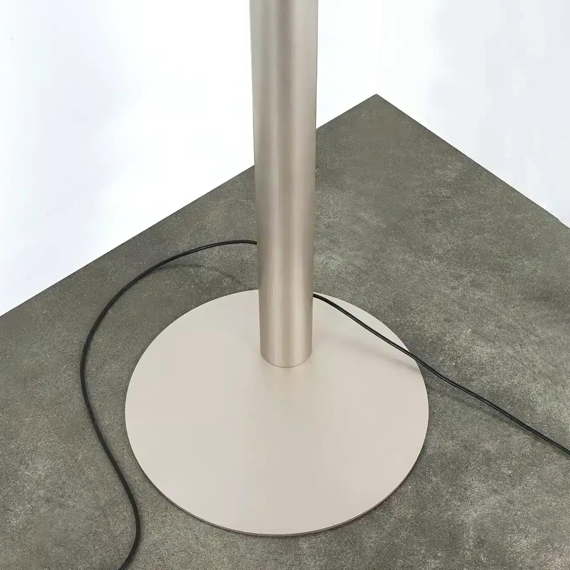 Close-up of a modern floor lamp base featuring a brushed metal cylindrical pole and a circular light gray base, resting on a dark gray concrete floor with a black electrical cord visible.