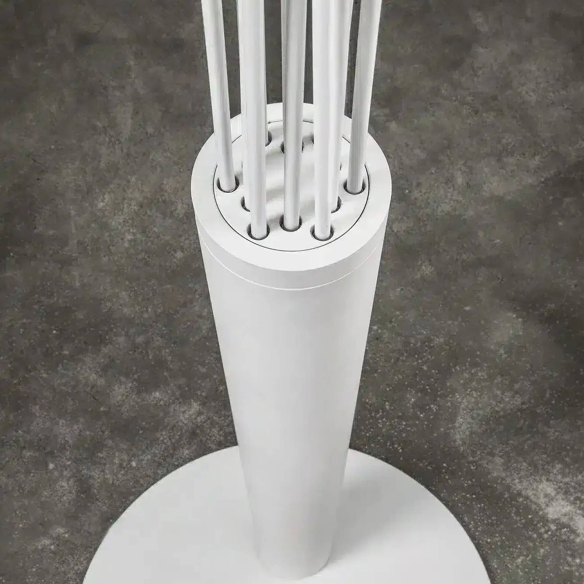 A white cylindrical object with a circular base, from which several thin white rods extend vertically upwards. It is set against a dark, textured concrete floor.