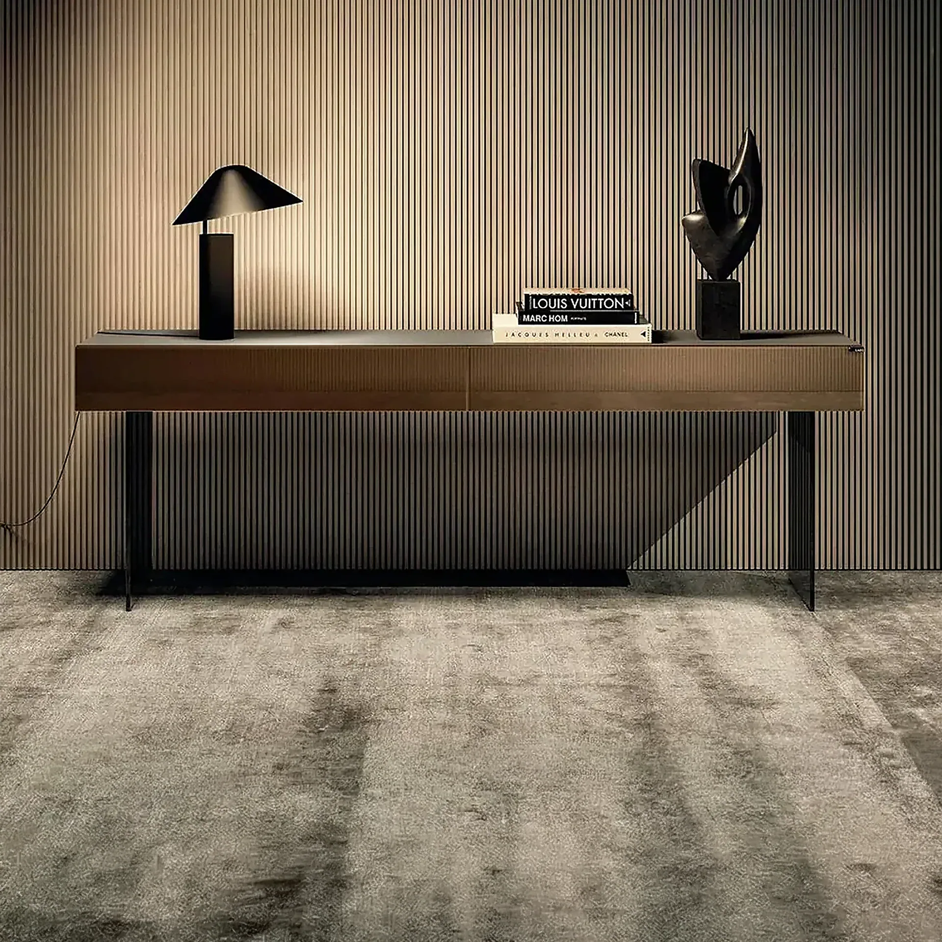 A modern console table with a black lamp, fashion books, and an abstract sculpture against a slatted wall and textured rug.