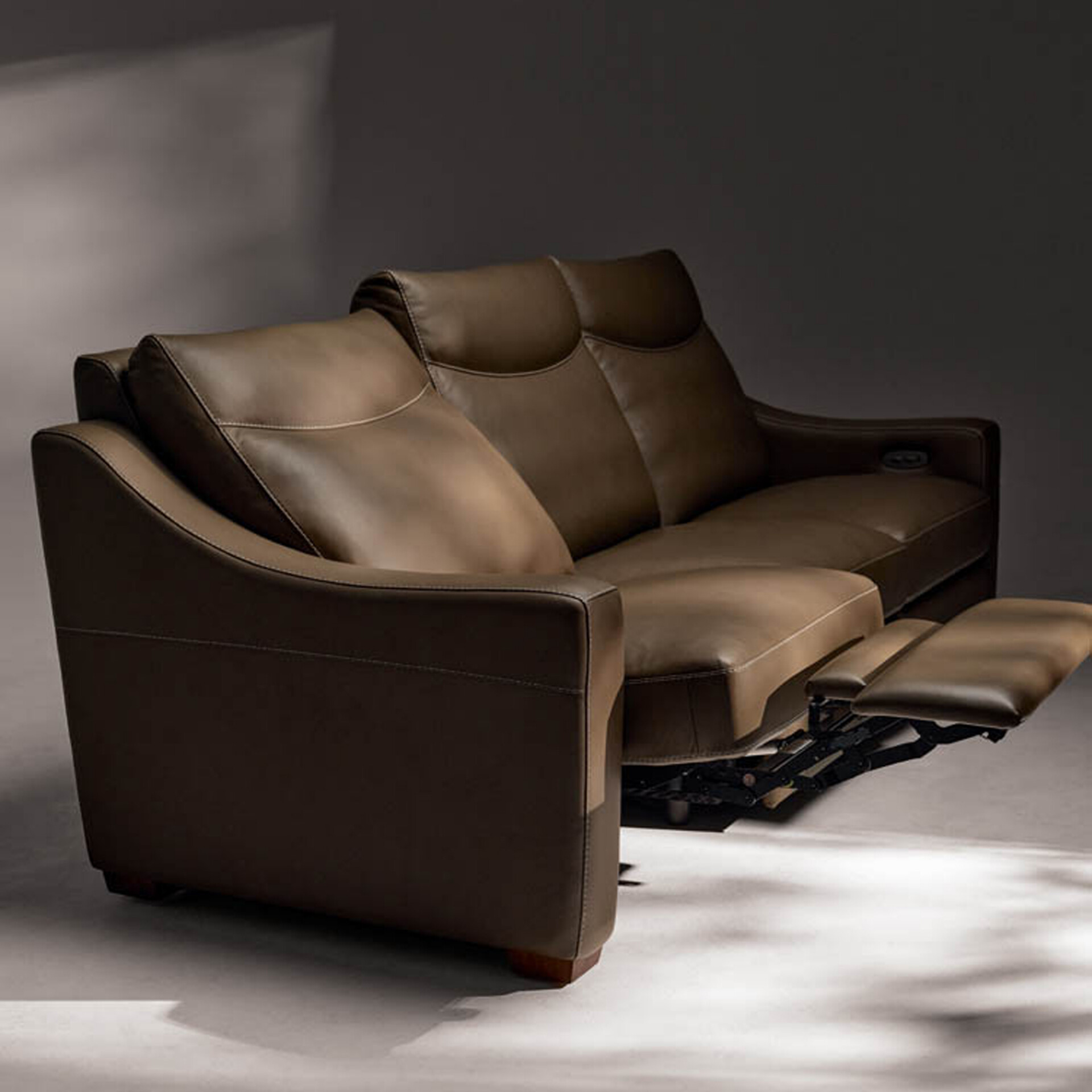 A brown leather two-seater reclining sofa with one footrest extended, against a dark gray background.