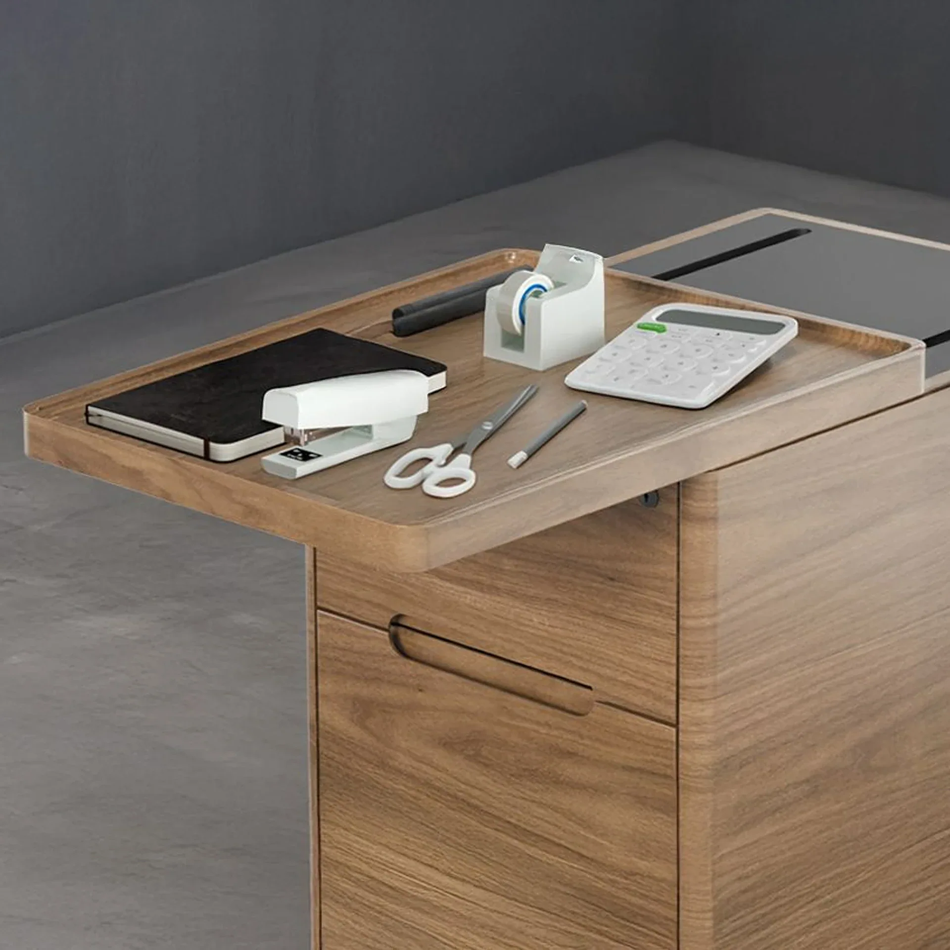 A wooden desk with a sliding tray holding a notebook, stapler, tape dispenser, calculator, scissors, and a pen.