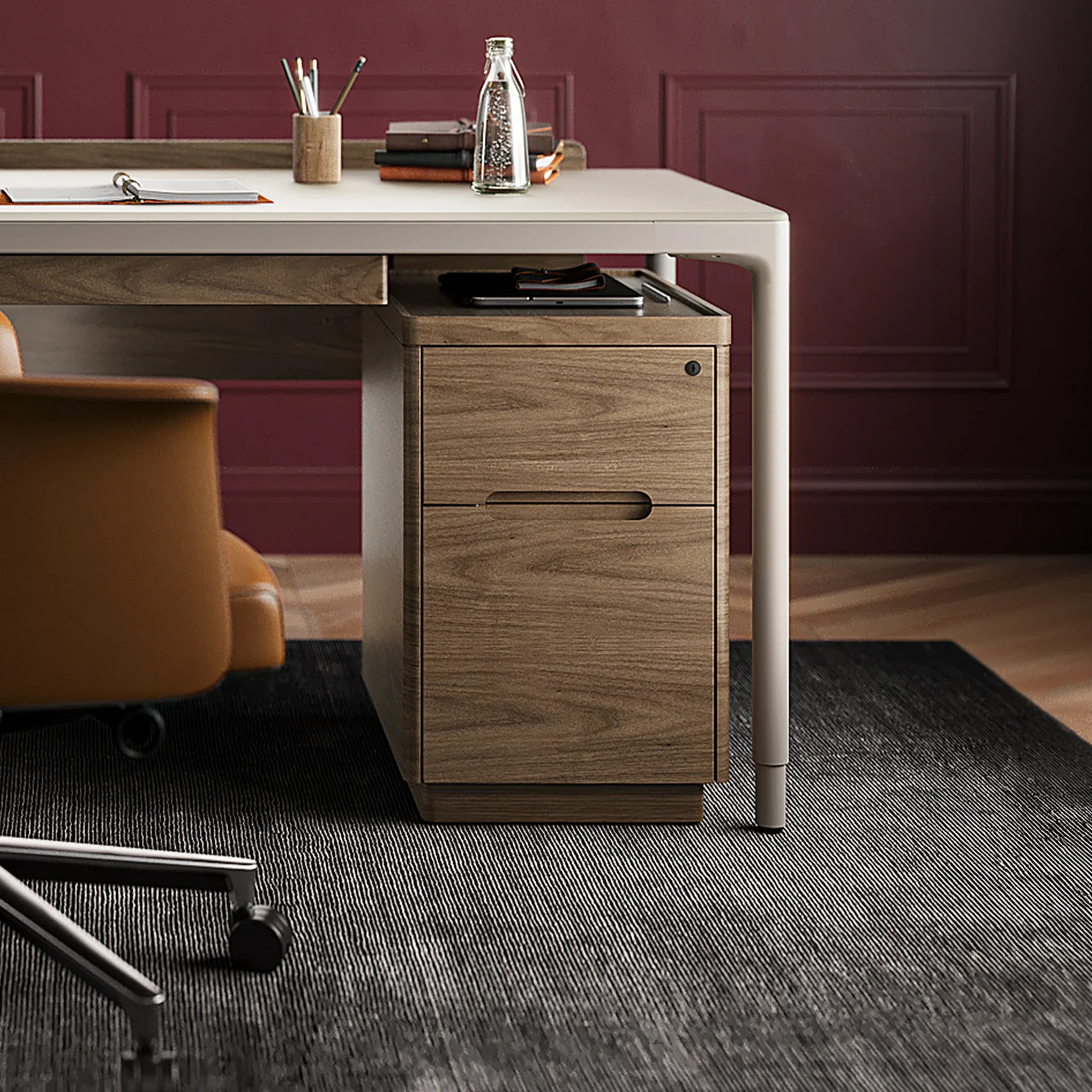 Modern office desk with a light top, wooden drawers, brown leather chair, and a dark red paneled wall.